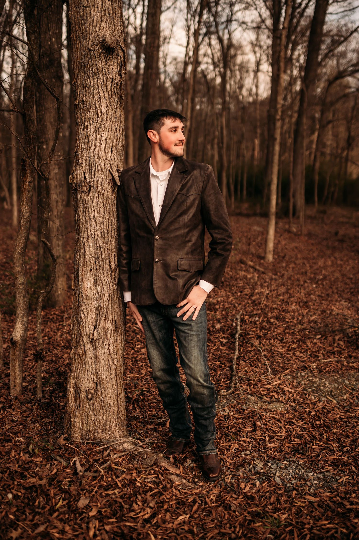 A groom leaning against a tree looking out to his left with fallen leaves under his feet.