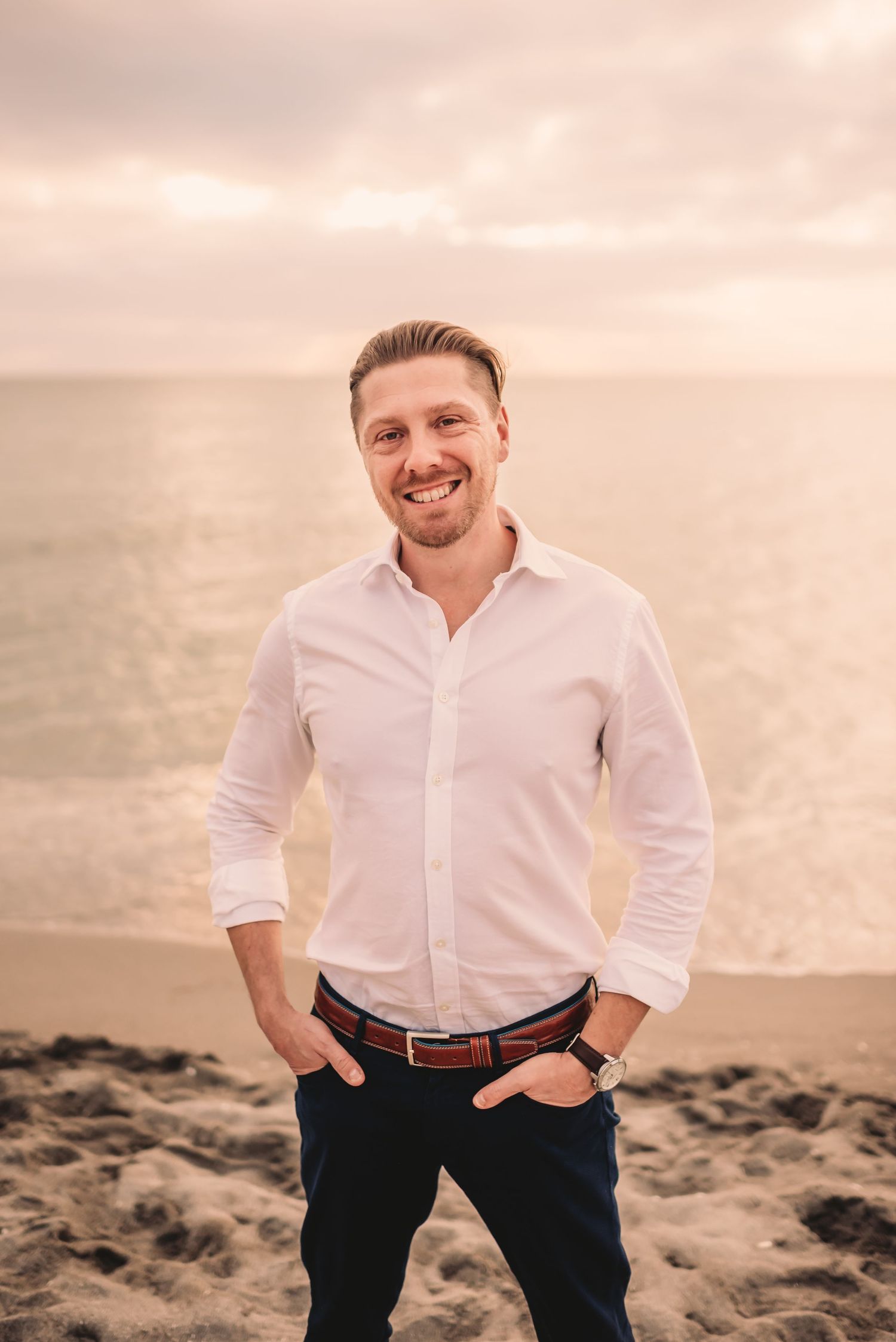 Groom stands with hands in pockets and smiling at camera on beach.