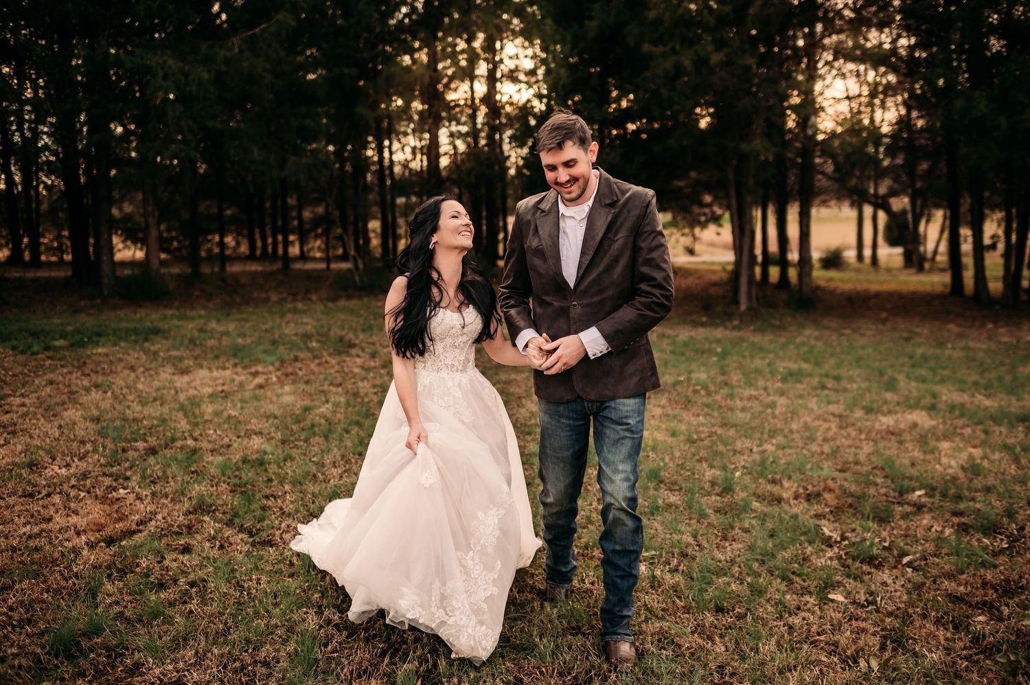 Newly married couple running and laughing int a field lined with trees while bride holds up her wedding dress.