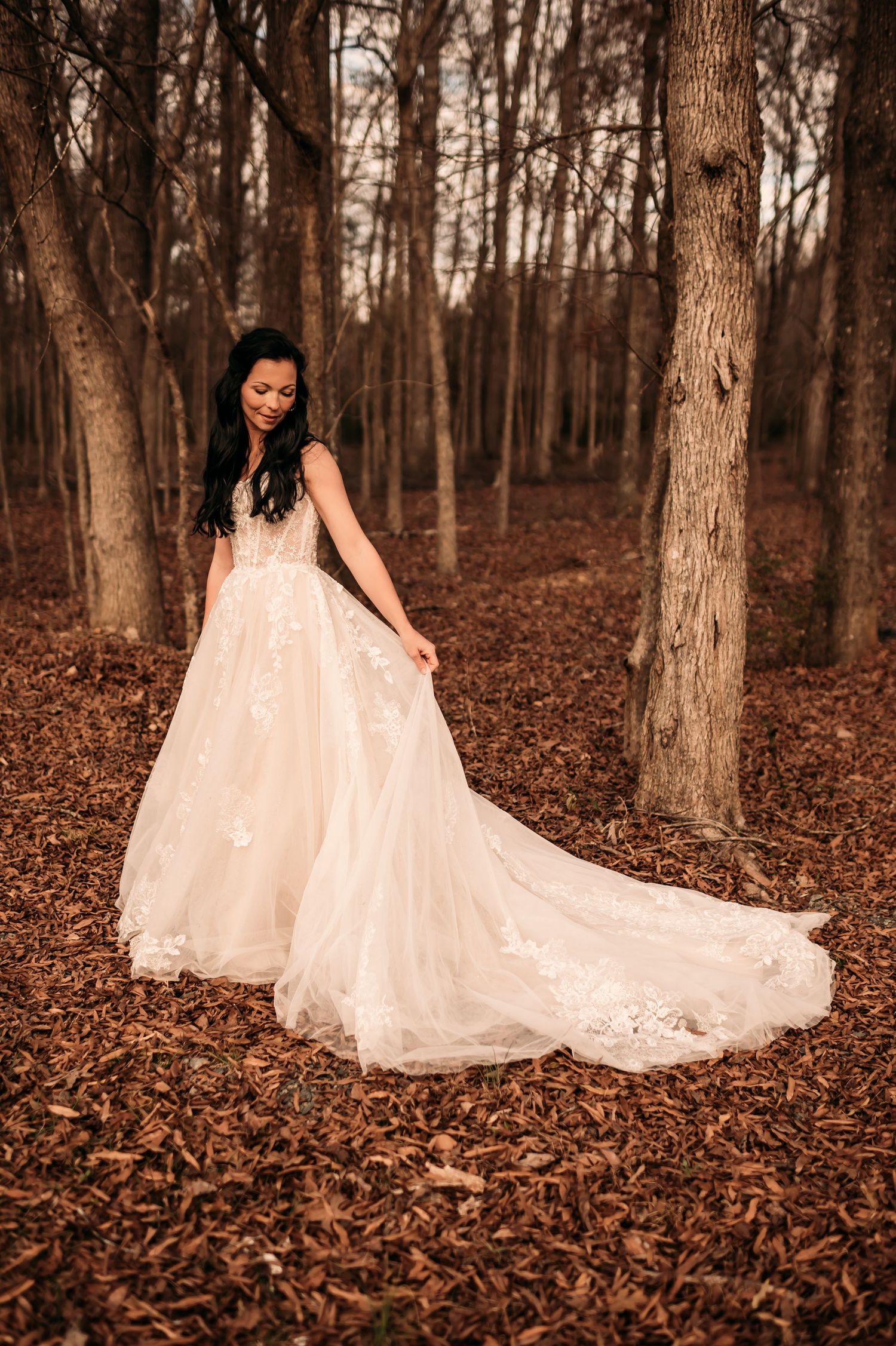 Bride holding her dress train to her left in front of tall trees.
