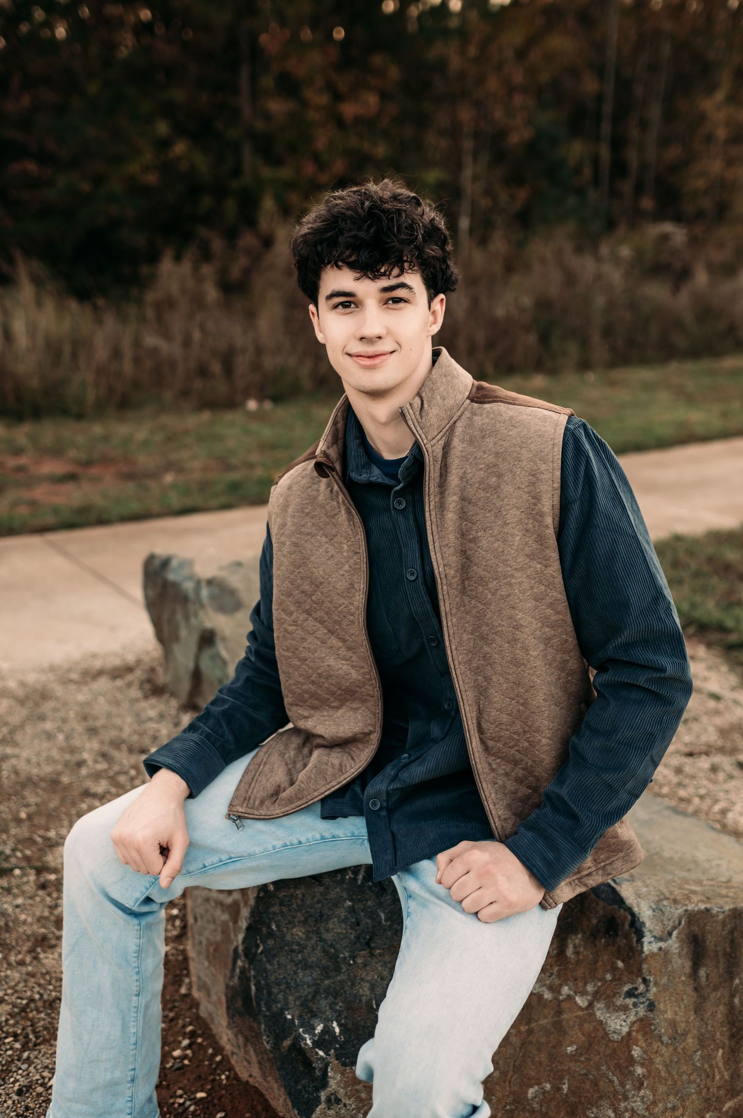 A teen boy sits on a rock and leans his right arm on his leg while smiling.