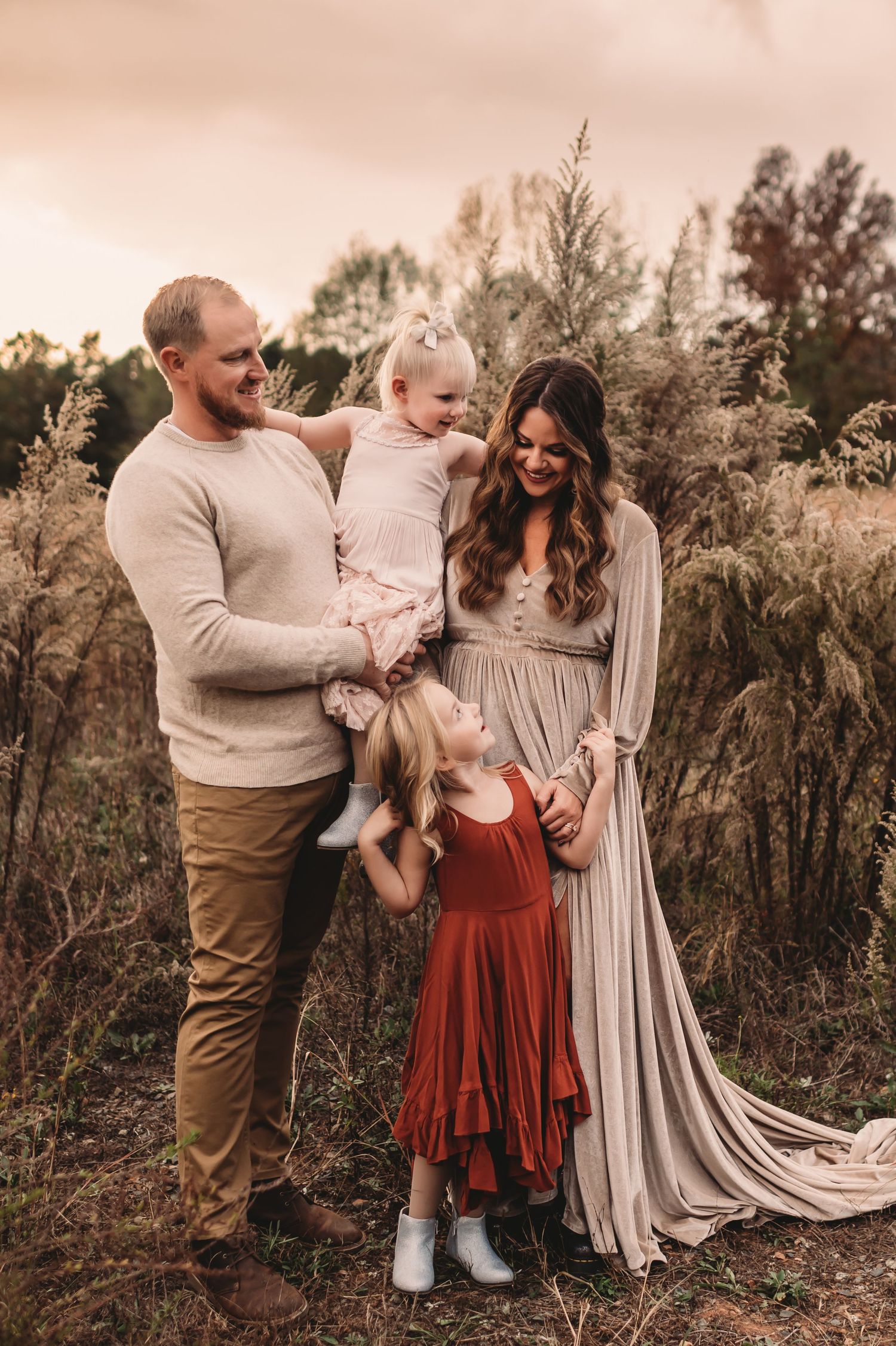 A mom and dad smiling while looking at their little girls in front of a tall grass field.