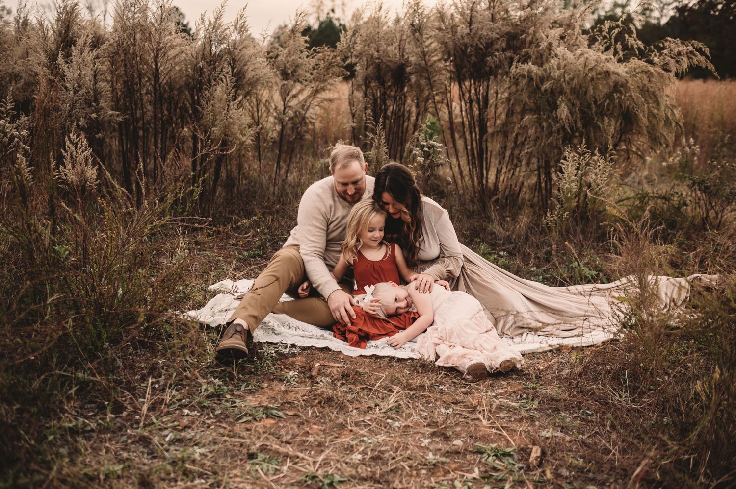 A mom and dad snuggling with their two daughters on a blanket surrounded by tall grass.