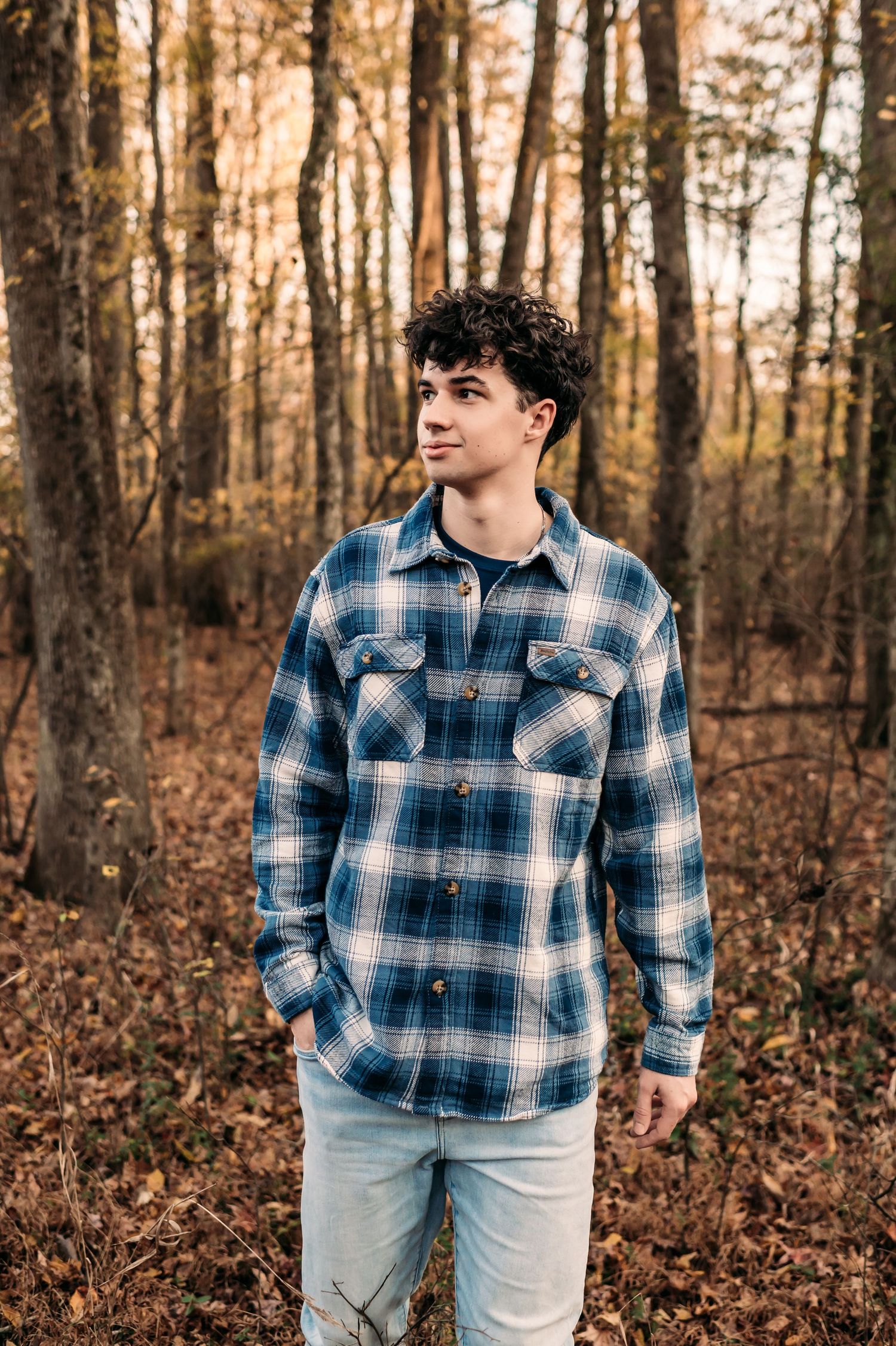 A senior boy walks through a golden forest in a plaid shirt with one hand in his picket.