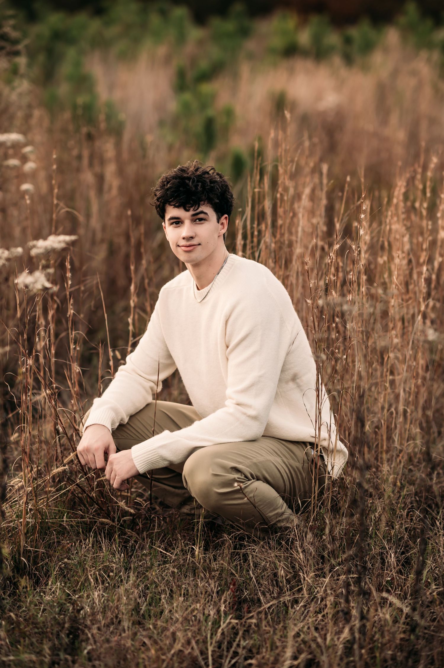 A senior boy crouches in a field of tall grass and smirks at the camera.