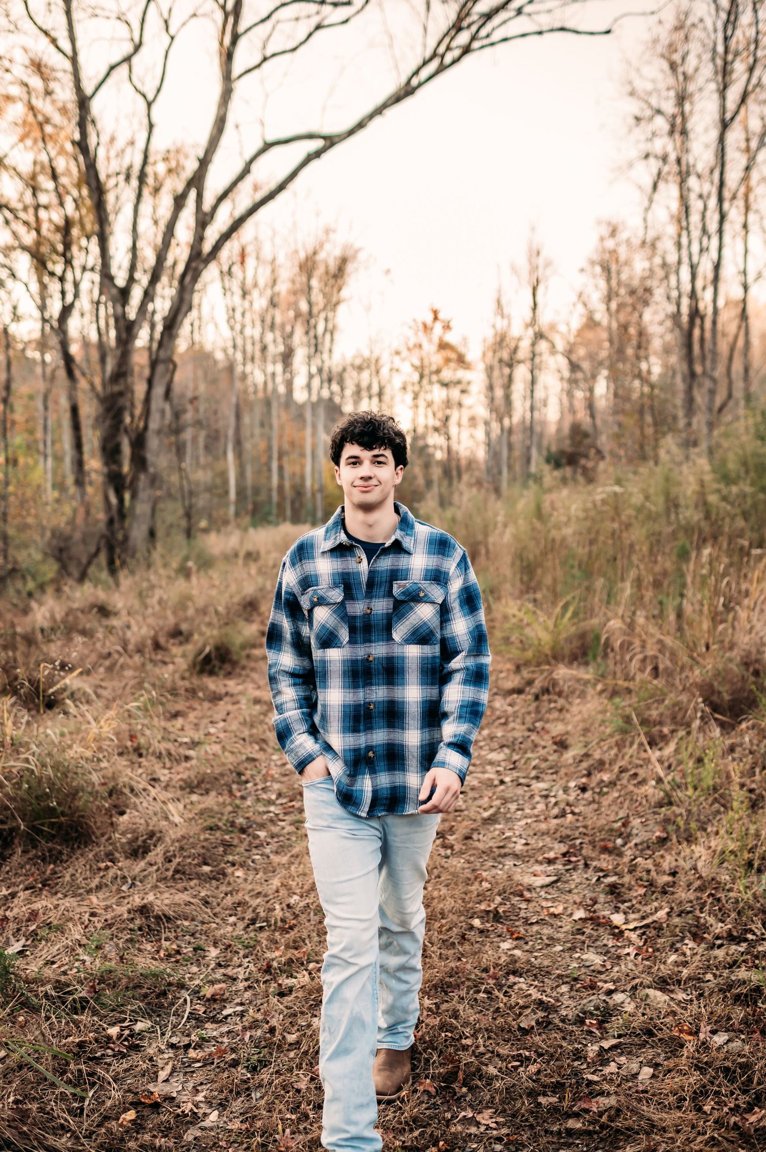 A senior boy in a plaid shirt walks in an open field in the forest and smirks.