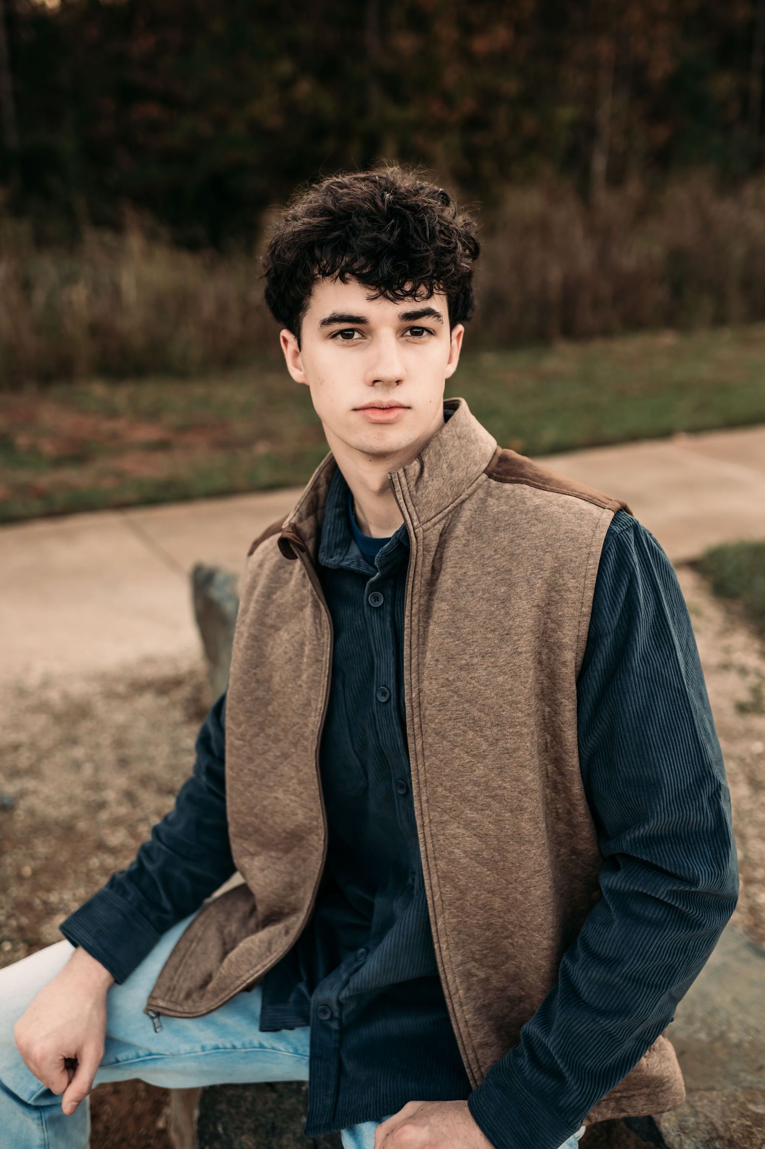 A teen sits on a large rock and wears a tan vest looking at a camera without smiling.