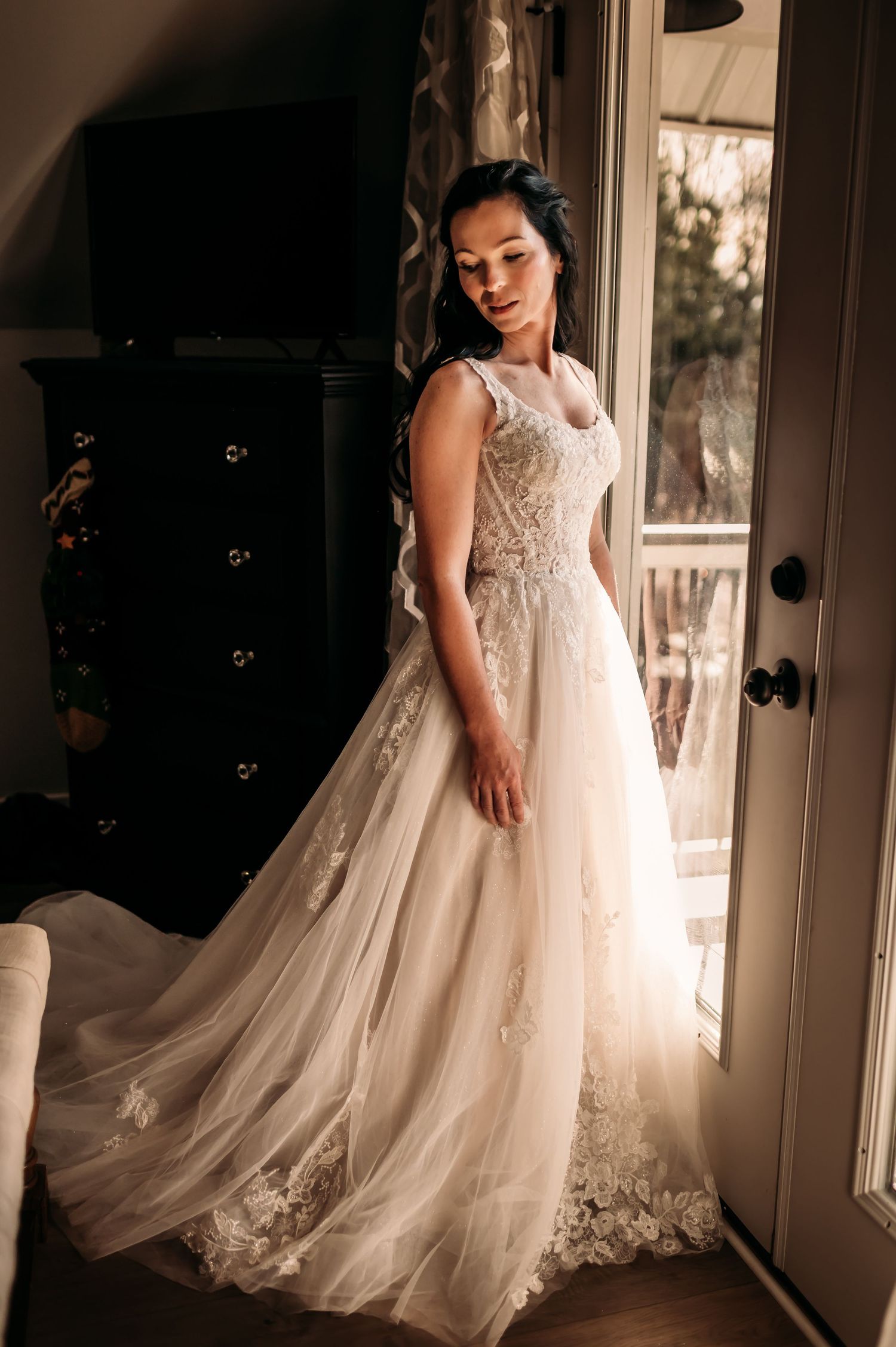 Woman in wedding dress by window looking down over her shoulder.