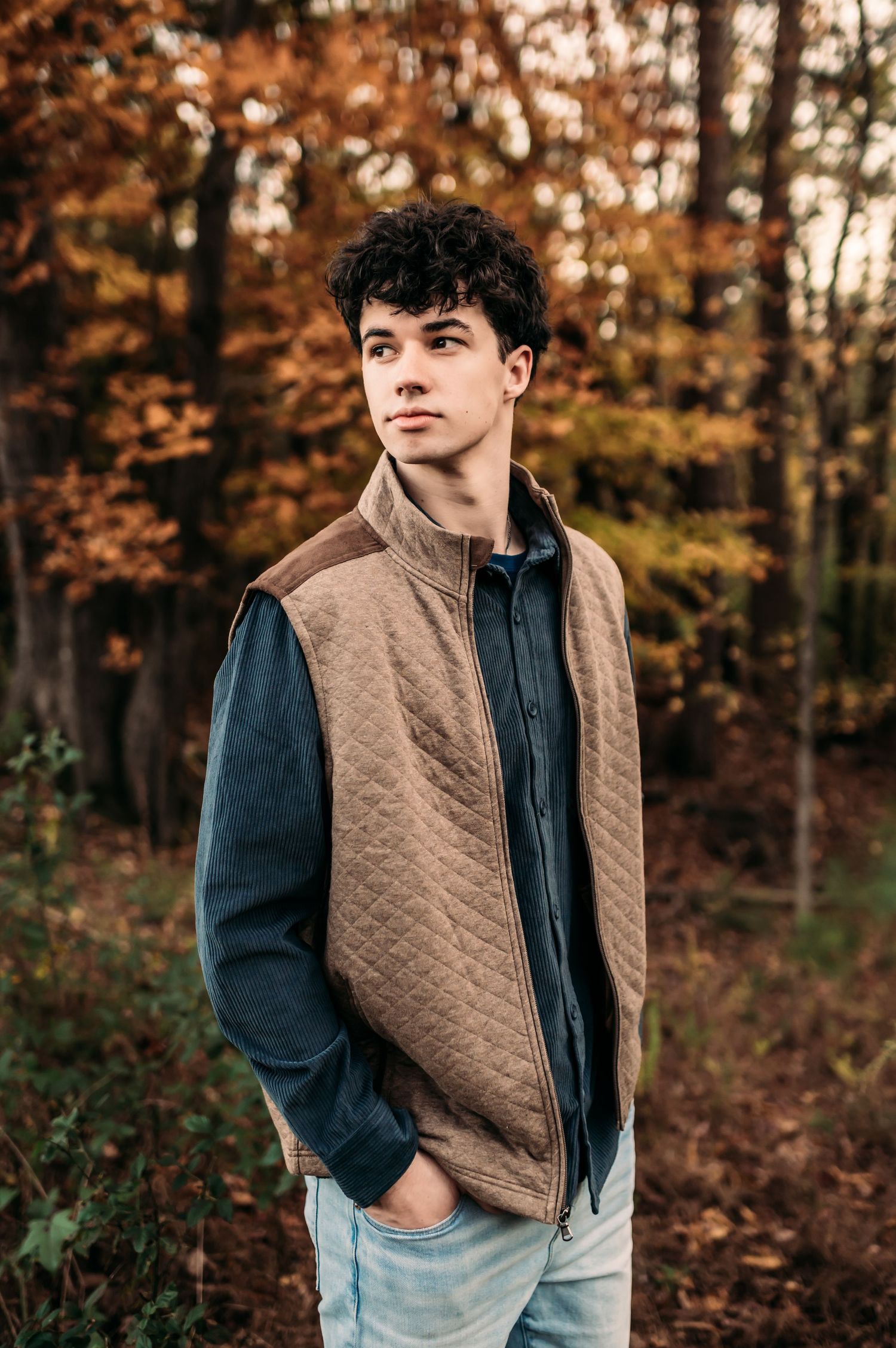 A teenager stands in a tan vest and dark long sleeved shirt looking out to his right.