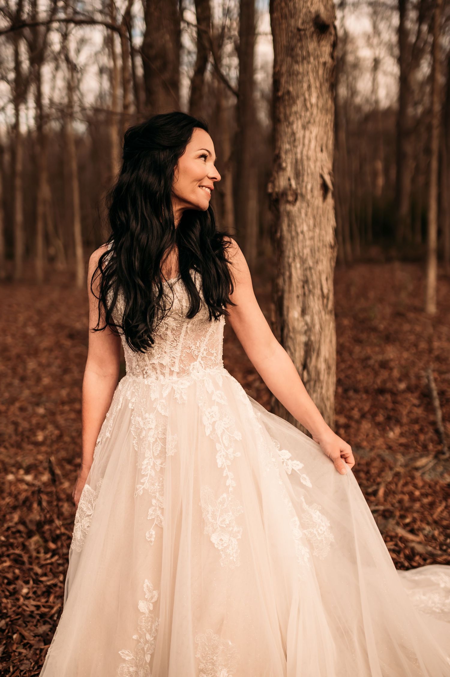 Bride holding her dress up in front of trees and looking over her shoulder with a soft smile.