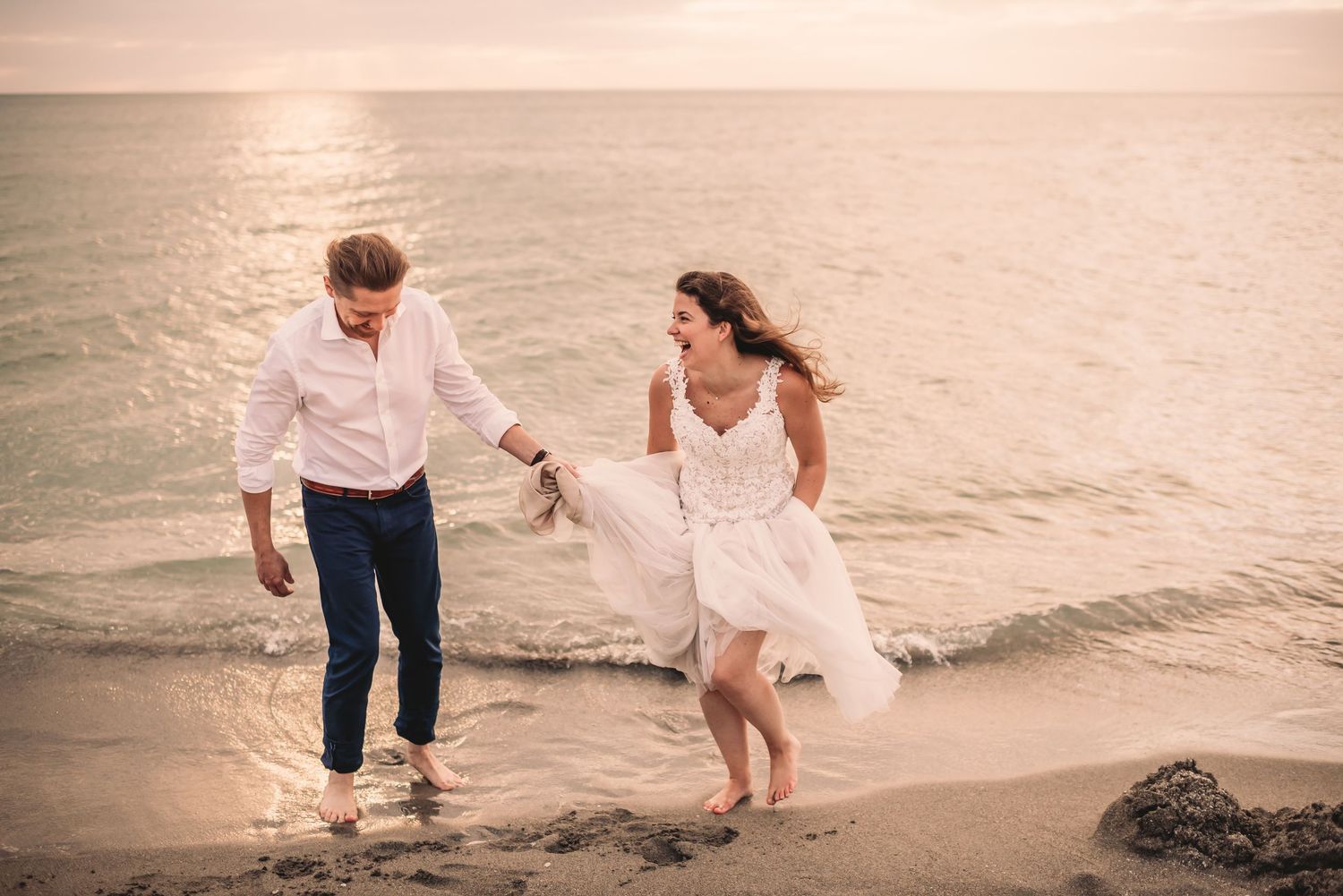 Bride and groom run from water on beach laughing together.