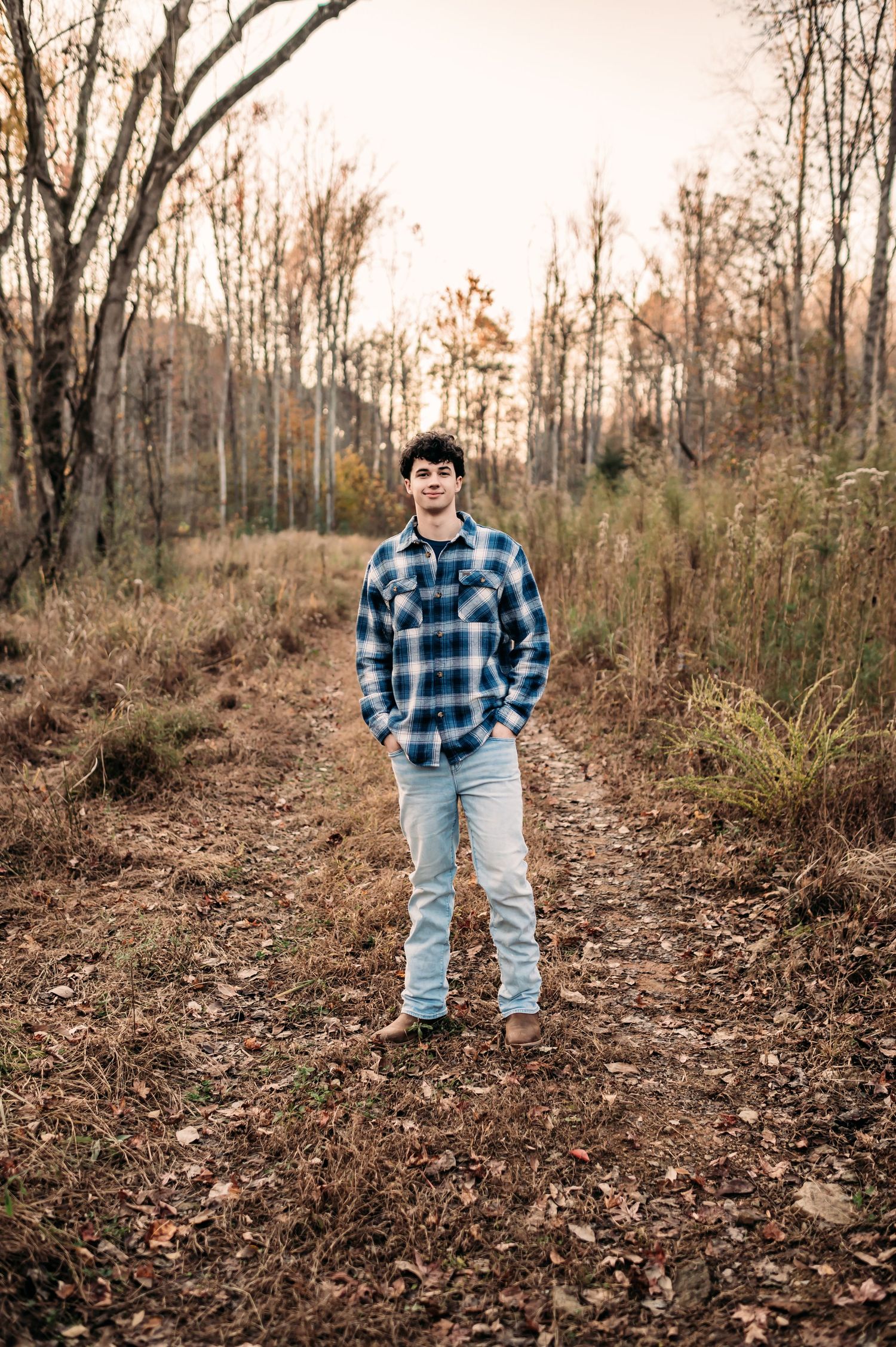 A teenage stands in an open area in a forest and smiles for the camera.