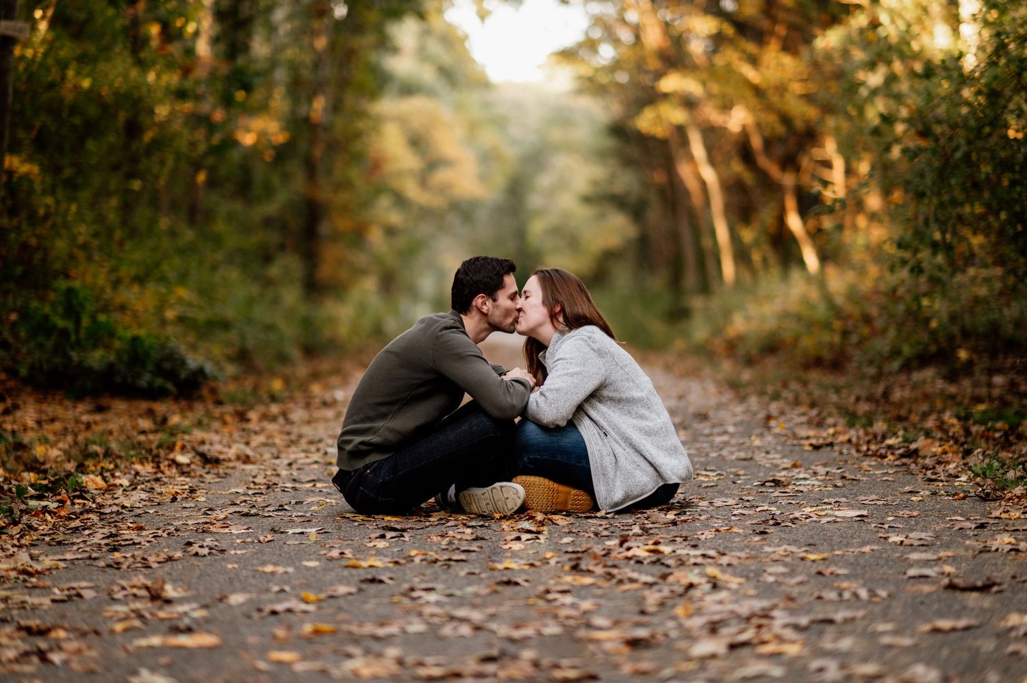 A romantic couple sits together on a wooded path covered in fallen autumn leaves.