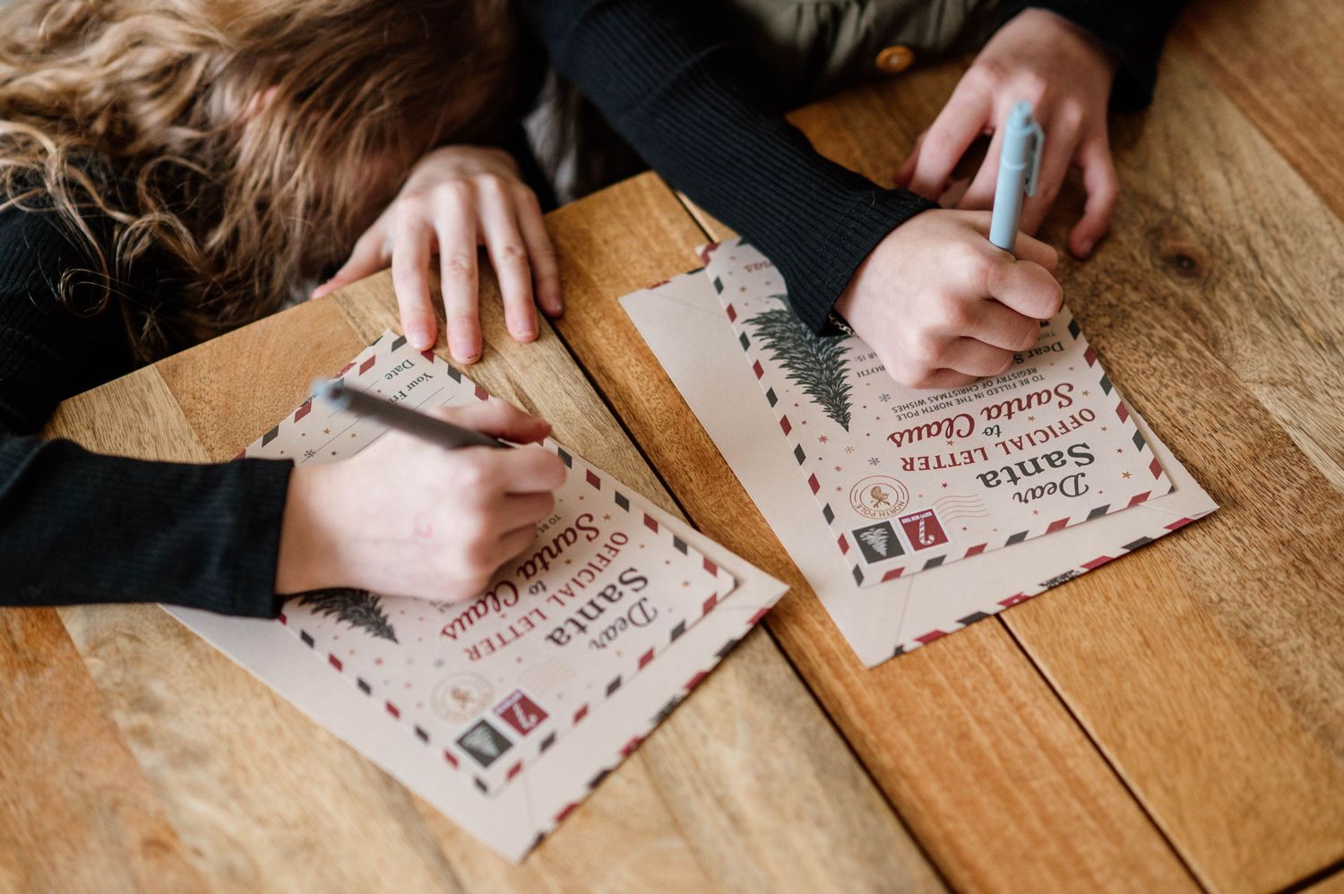 Two children writing letters to Santa Claus on decorated envelopes at a wooden table during the Christmas holiday season.