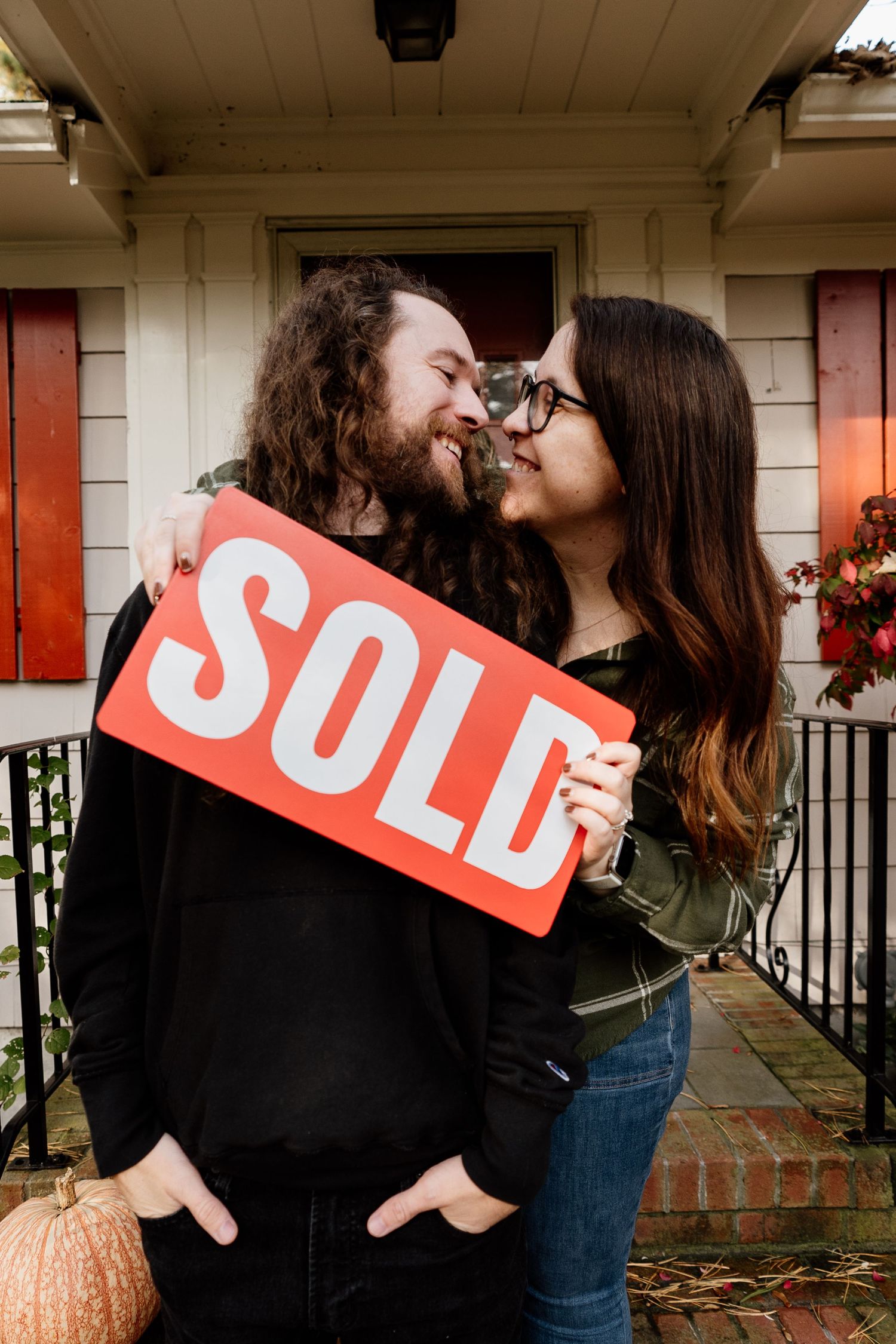 A couple holds a red SOLD sign while sharing a romantic moment in front of a house with a pumpkin on the porch.