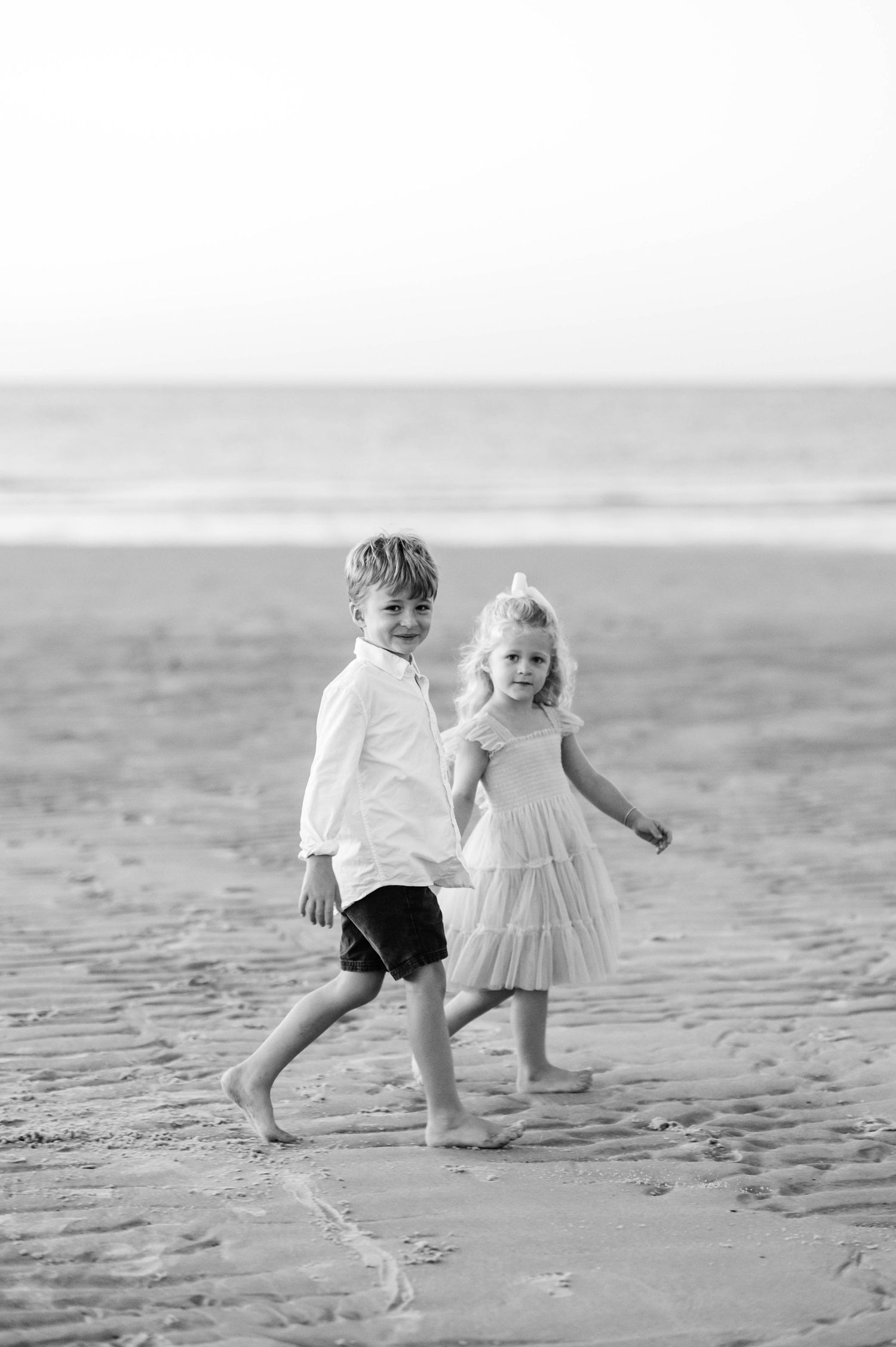Two small children playing and running together on a sandy beach in black and white.
