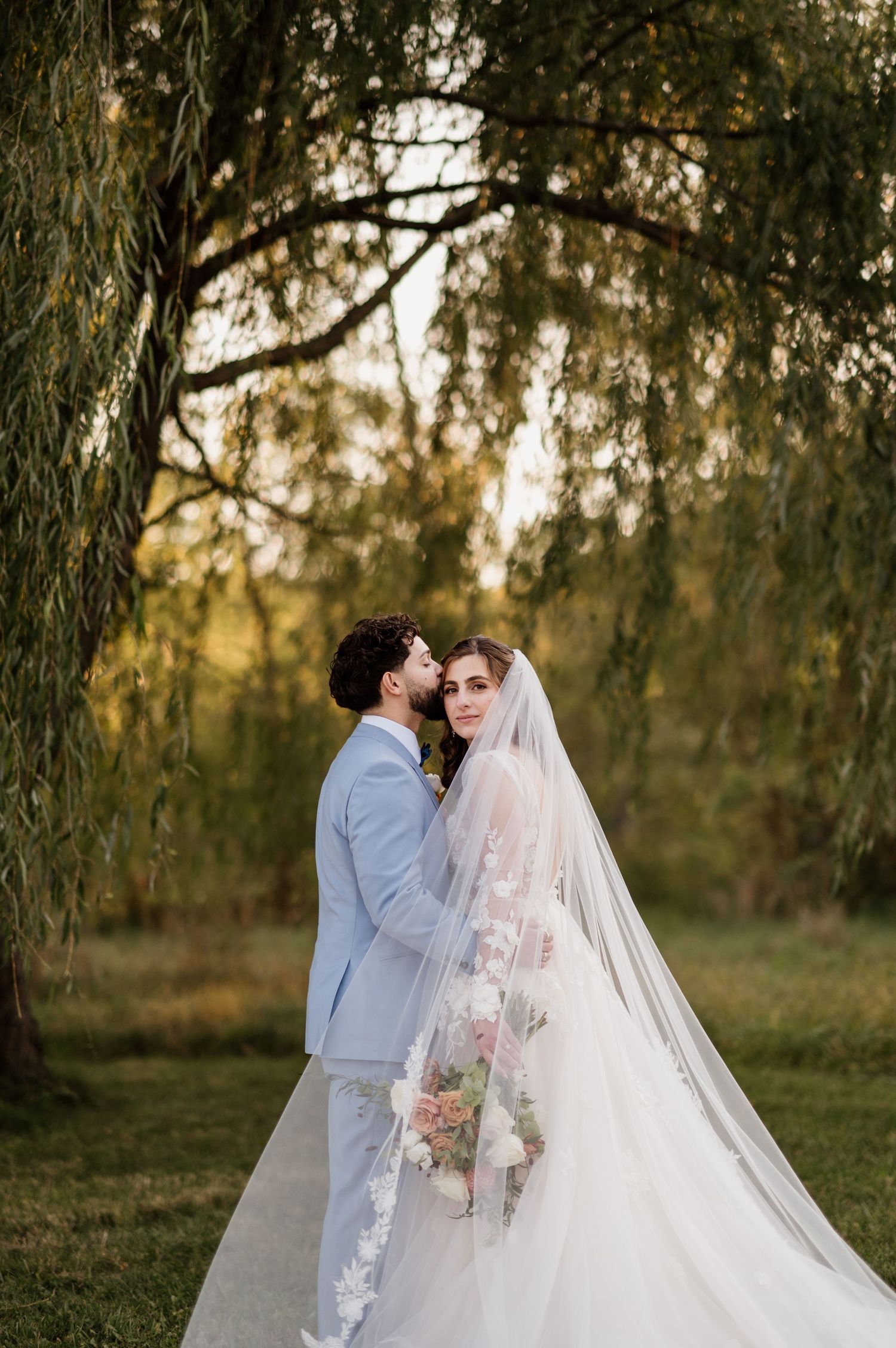 Wedding couple shares tender moments under willow trees during golden hour.