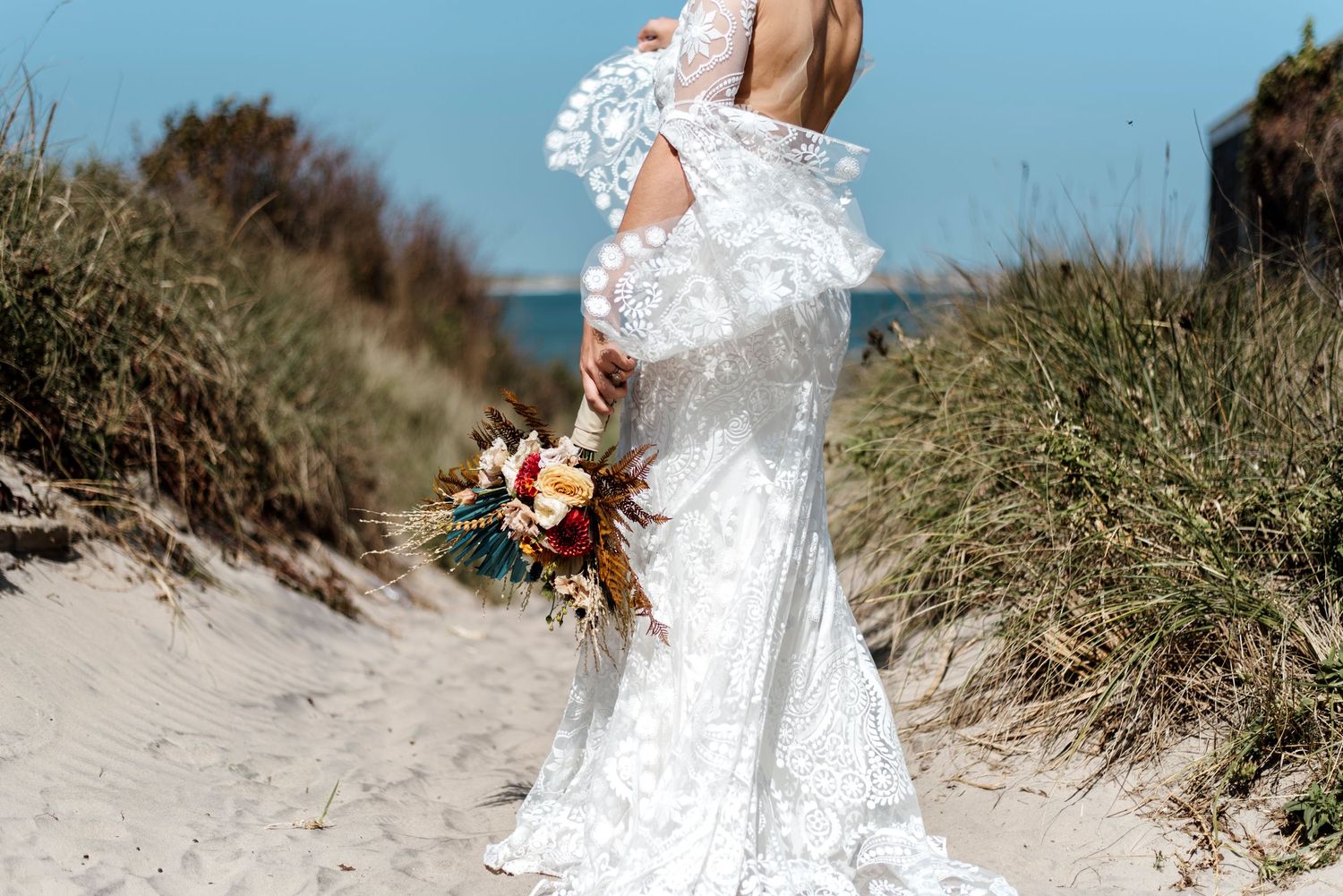Wedding dress train flows in the wind against coastal dunes and beach grass.