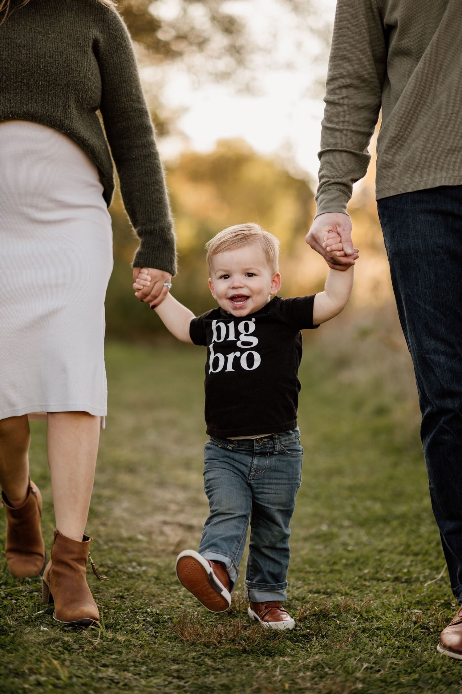 Small child wearing a shirt with text walking between two adults on grass at sunset.