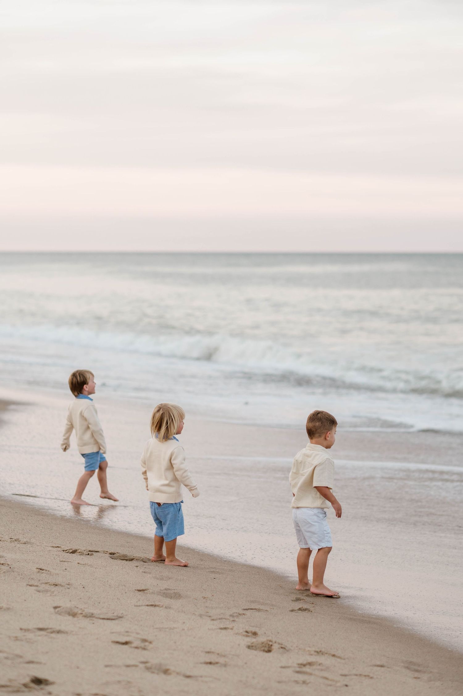 Three small children run and play together on a sandy beach at sunset.