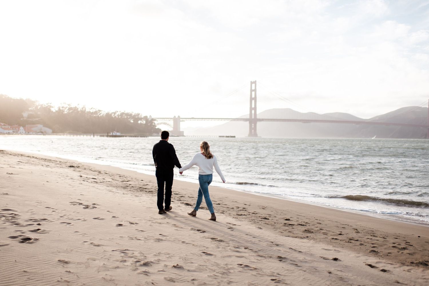 A series of silhouettes walk along a foggy beach with the Golden Gate Bridge visible in the distance.