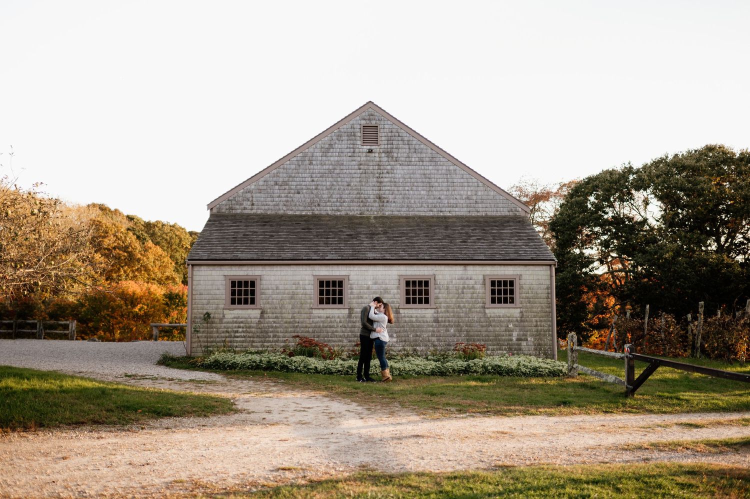 A couple stands in front of a historic grey stone barn on an autumn day.