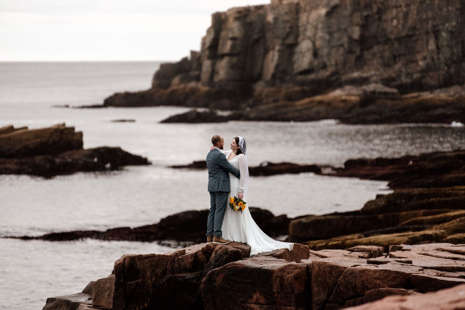 A couple poses on dramatic coastal rocks during their wedding photoshoot with ocean waves crashing in the background.