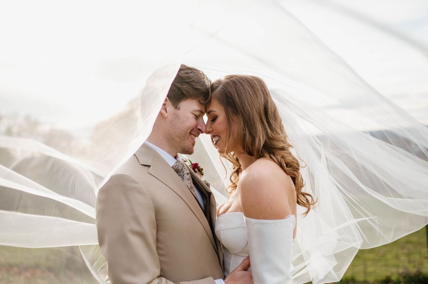 A romantic wedding couple embraces under a flowing white veil in a natural outdoor setting.
