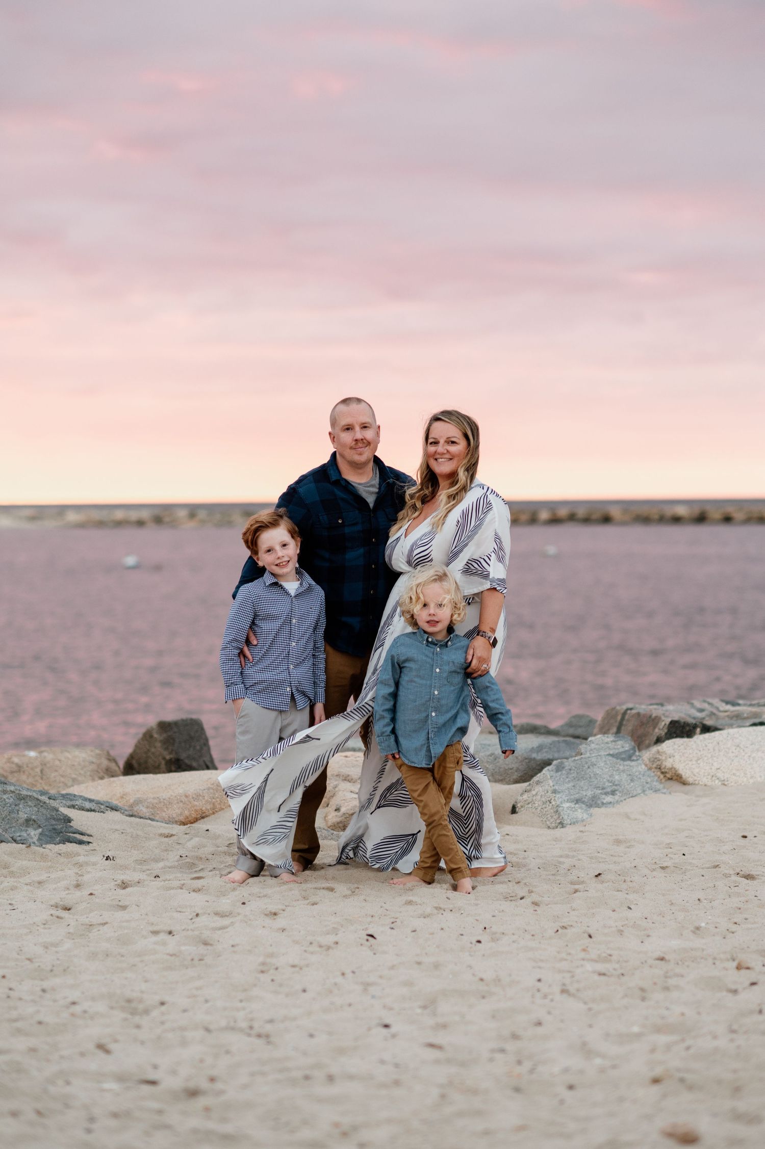 Family portrait on rocky beach during sunset with pink sky in background.