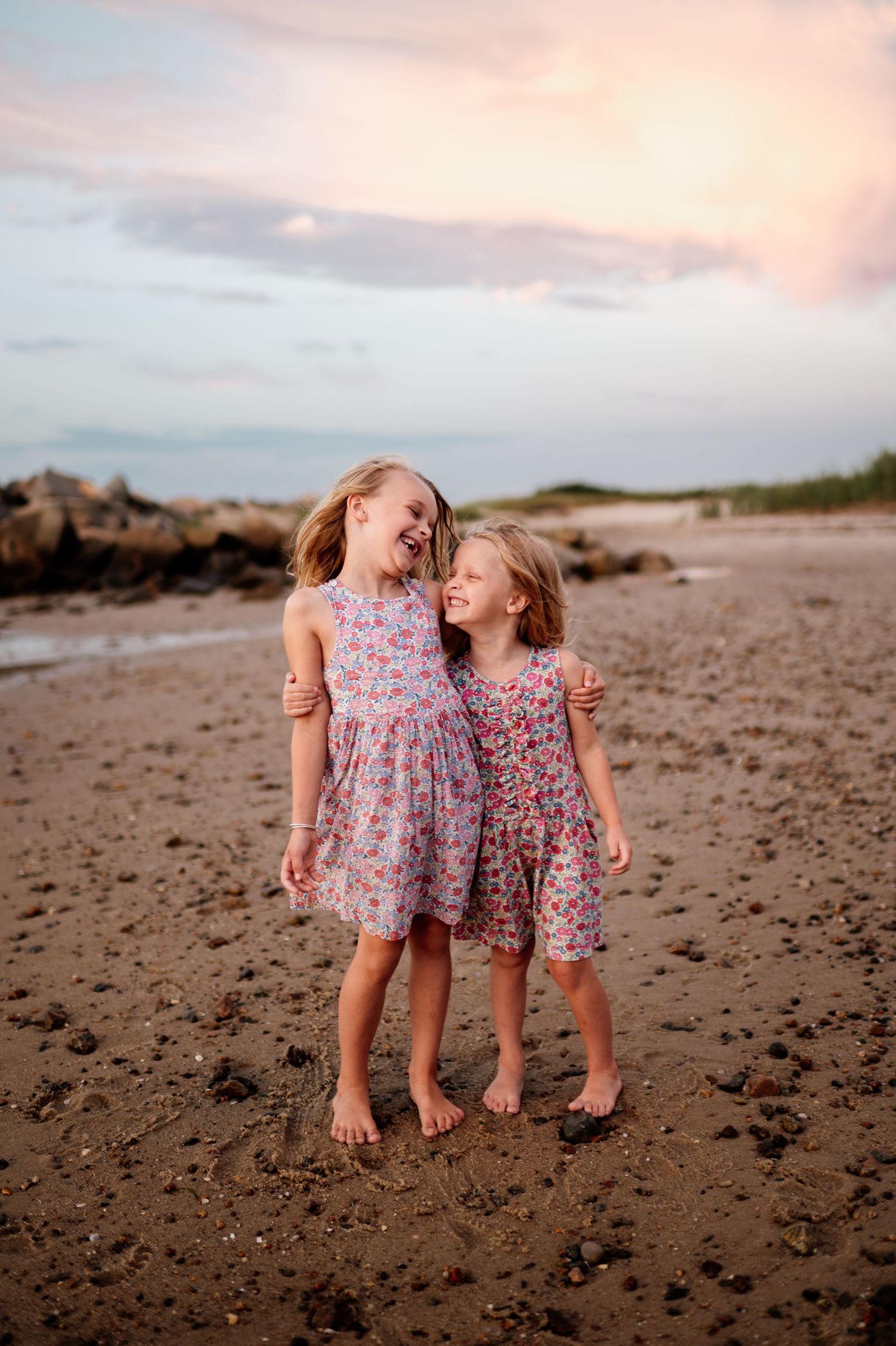 Two young girls in floral dresses play and laugh together on a sandy beach at dusk.