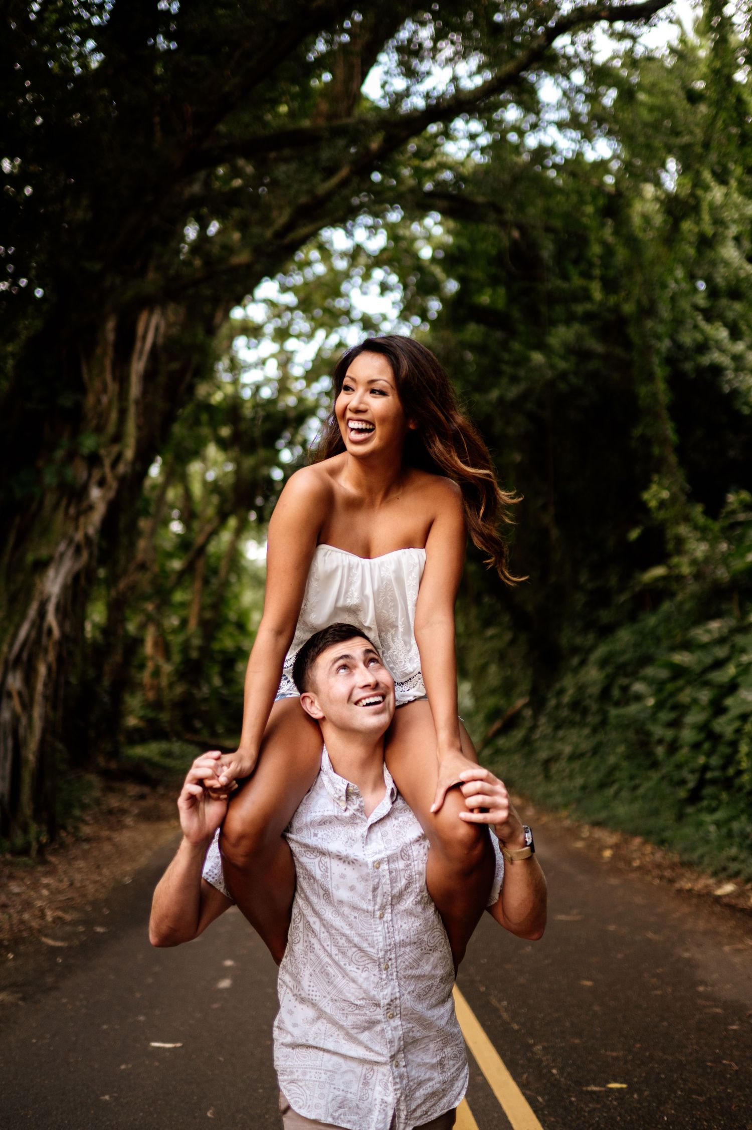Playful couple laughs together on a tree-lined path during outdoor photoshoot.
