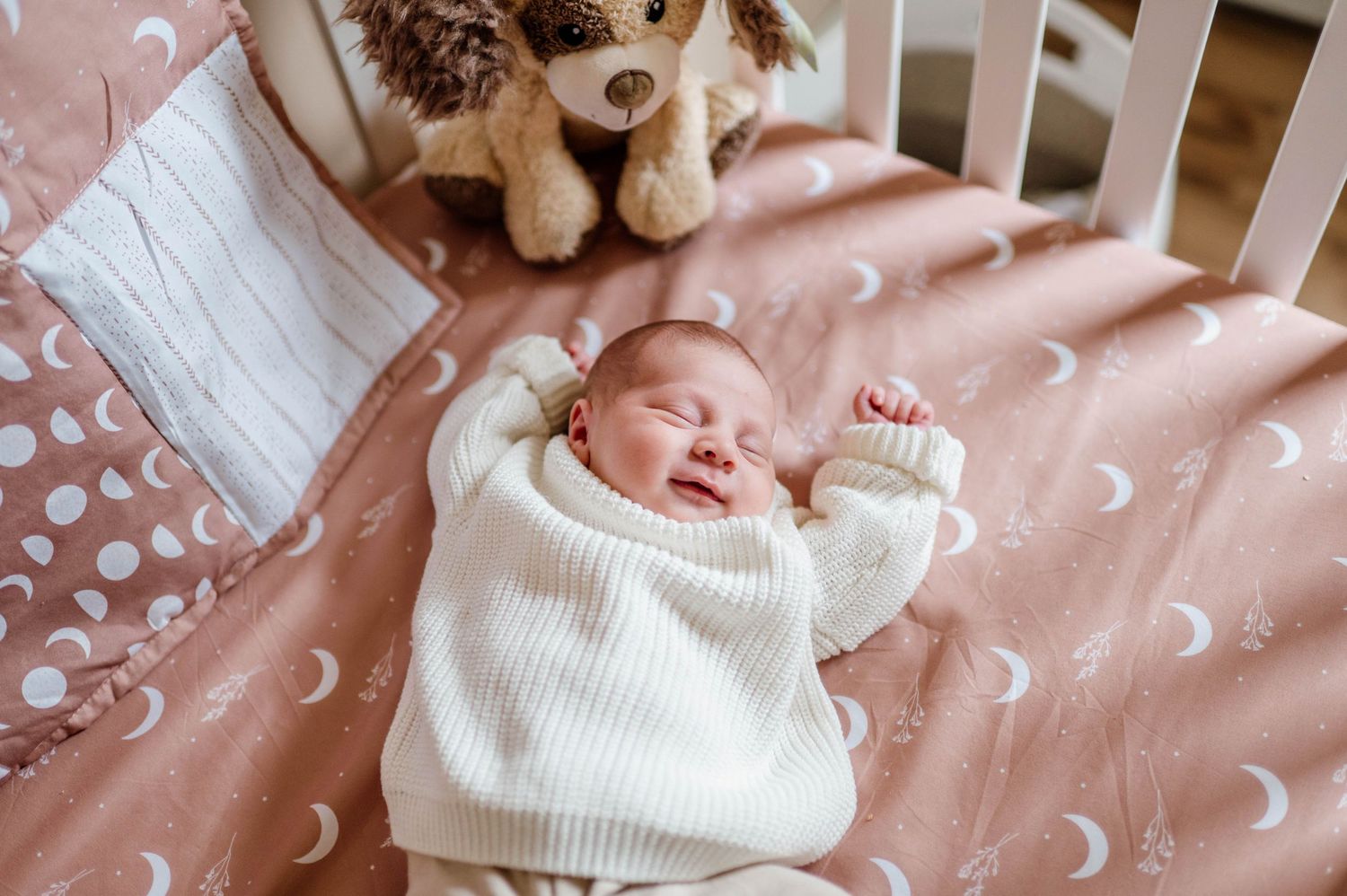 A sleeping infant in a white sweater lies on pink polka dot bedding next to a plush toy.