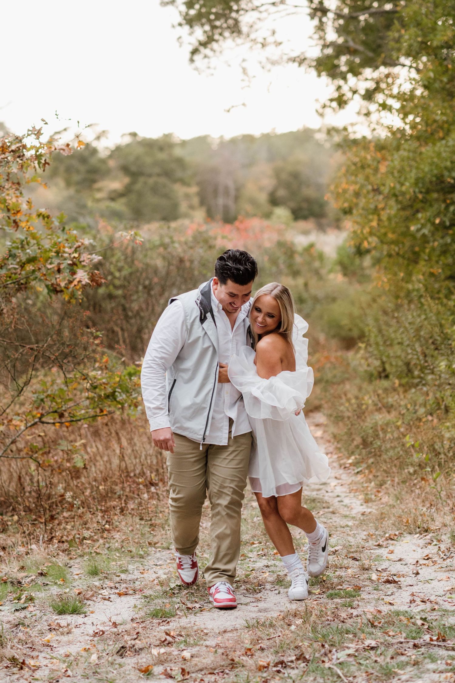 A couple shares an intimate moment while walking on a leaf-covered autumn path surrounded by trees.