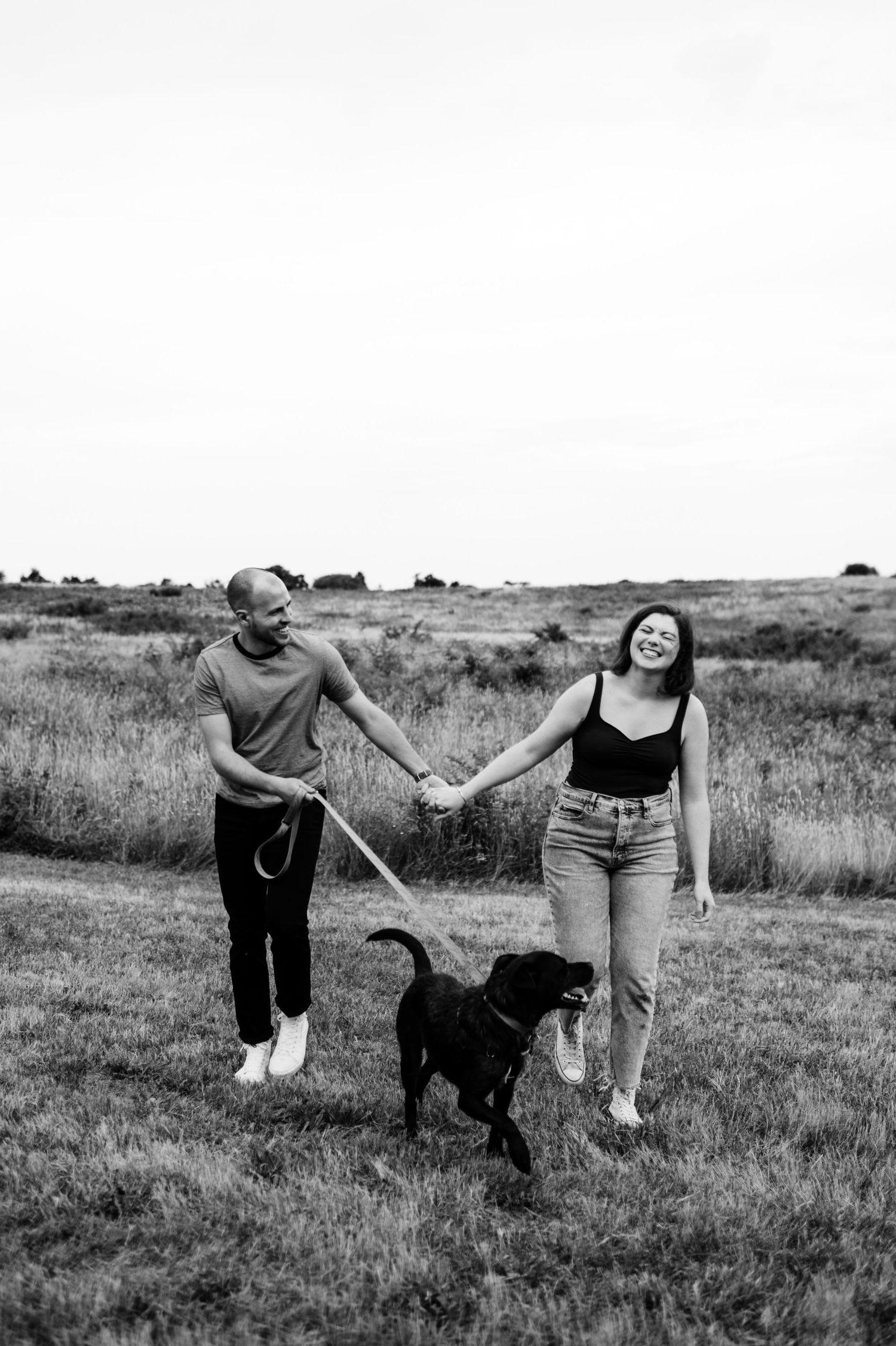 Black and white photo of a couple walking their dog through a grassy field.