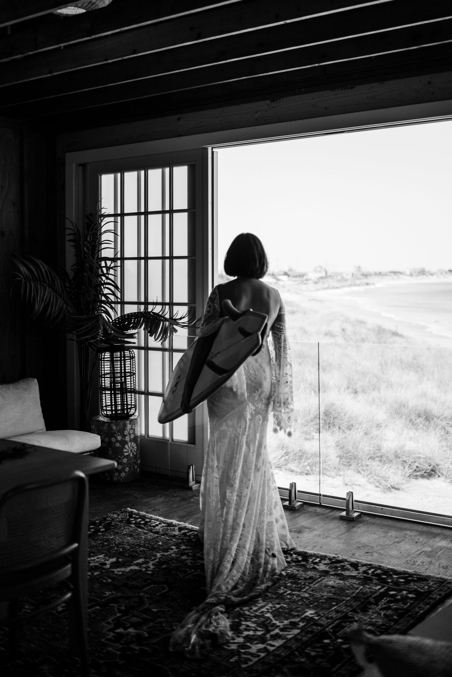 Silhouette of bride in flowing dress looking out large windows at coastal view.