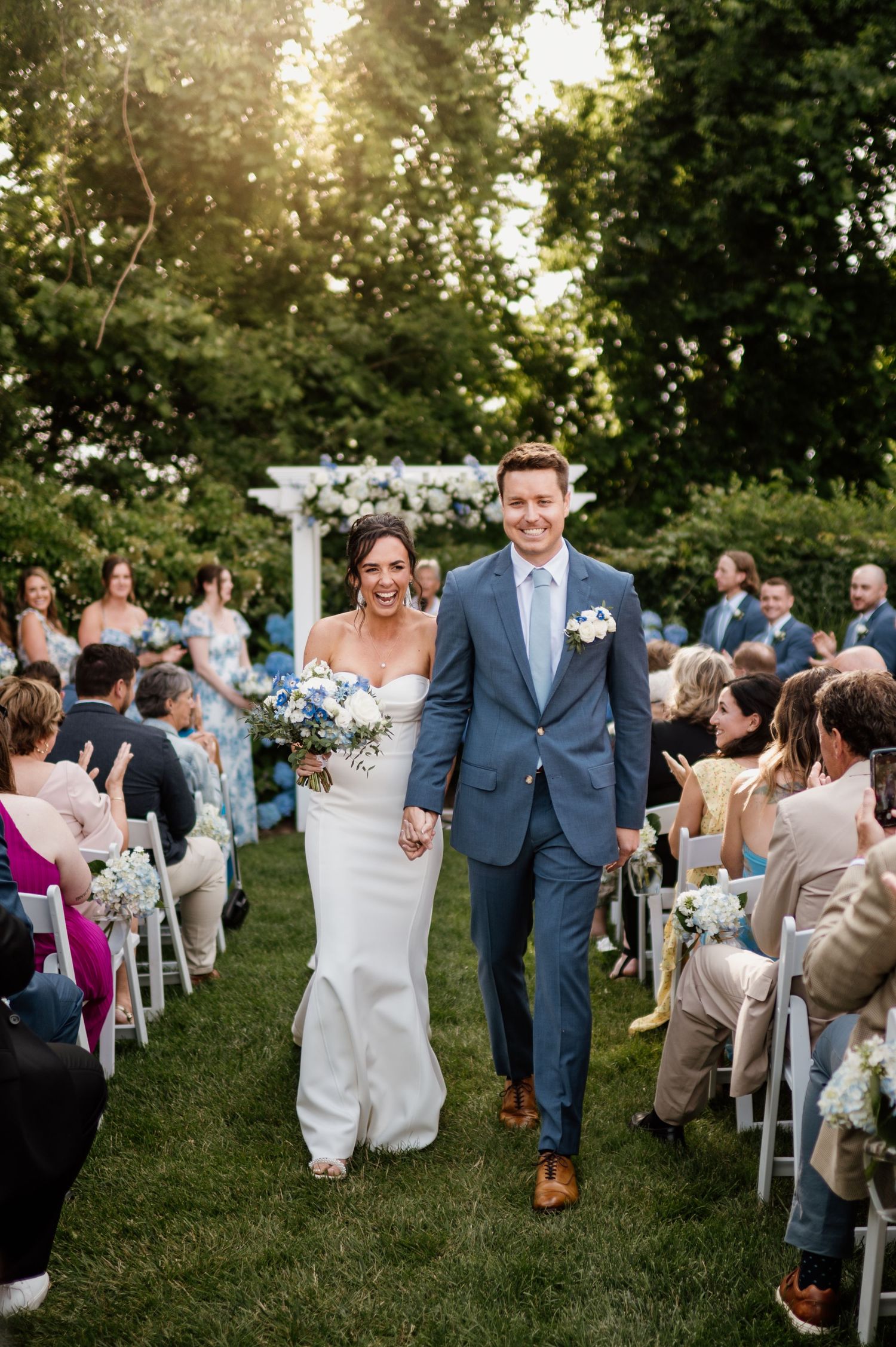 Newlyweds walk down the aisle between rows of seated guests at an outdoor garden wedding ceremony.