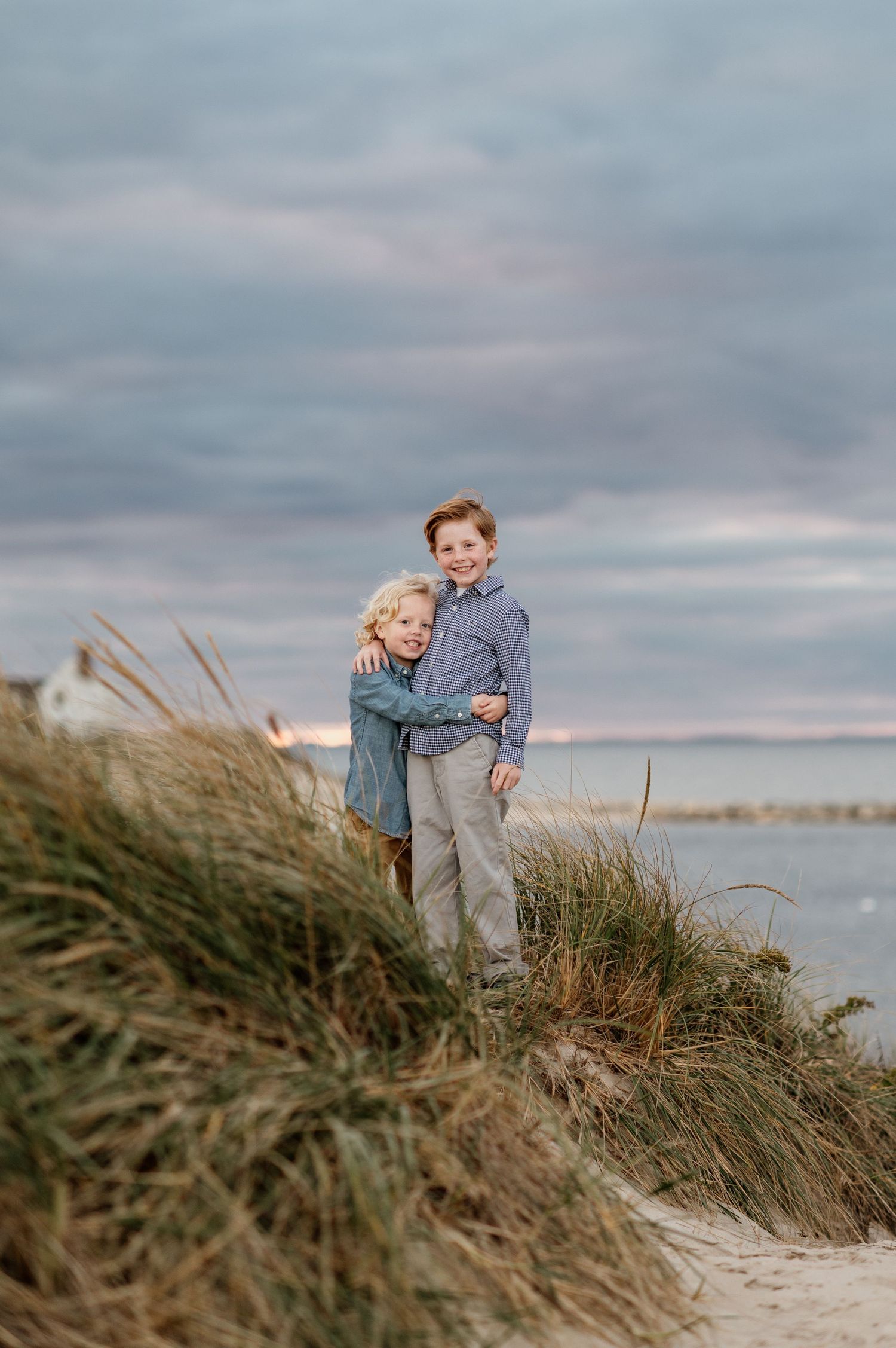 Two figures stand among windswept beach grass on coastal dunes under a cloudy sky.