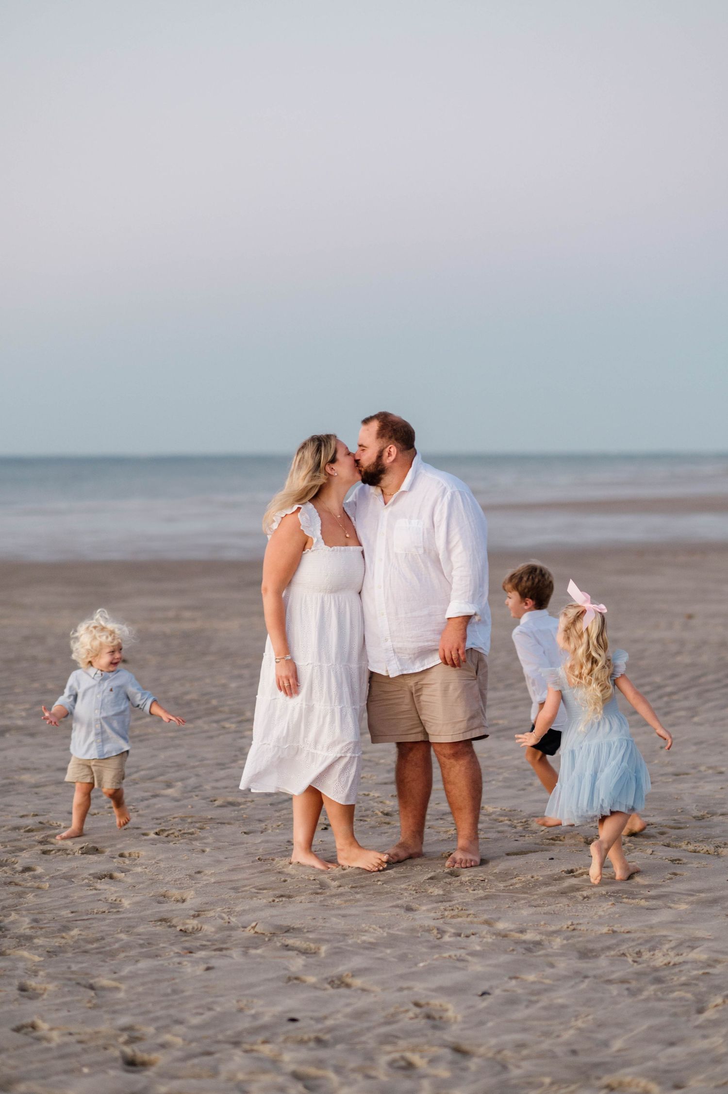 Family in coordinated white and beige outfits walking along beach at sunset.