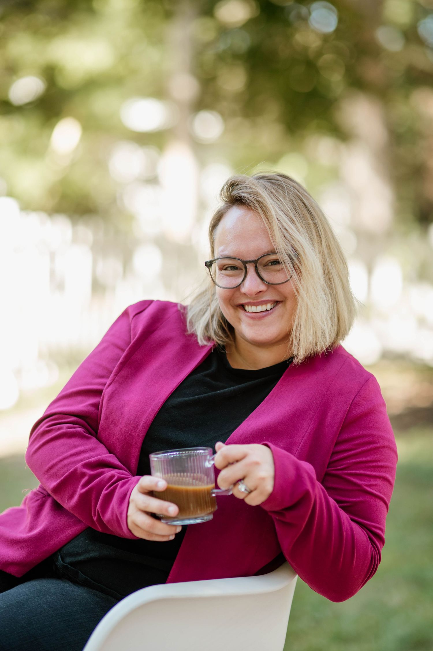 A person in a bright pink blazer and black top smiles while holding a beverage outdoors.