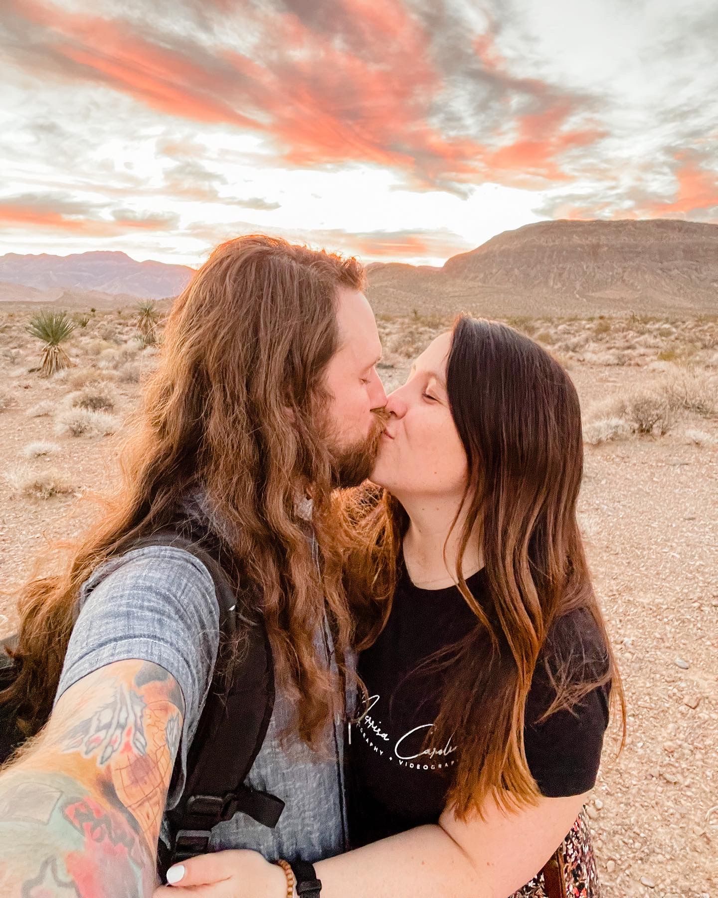 A couple shares a romantic kiss in the desert during sunset with warm orange skies and mountains in the background.