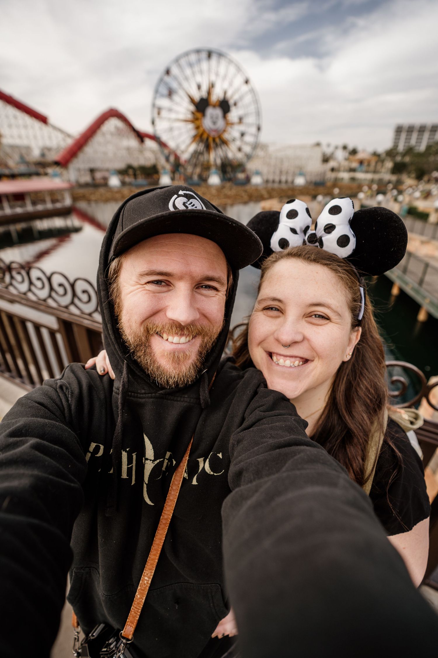 A couple wearing black clothing takes selfies together at an amusement park with carnival rides in the background.