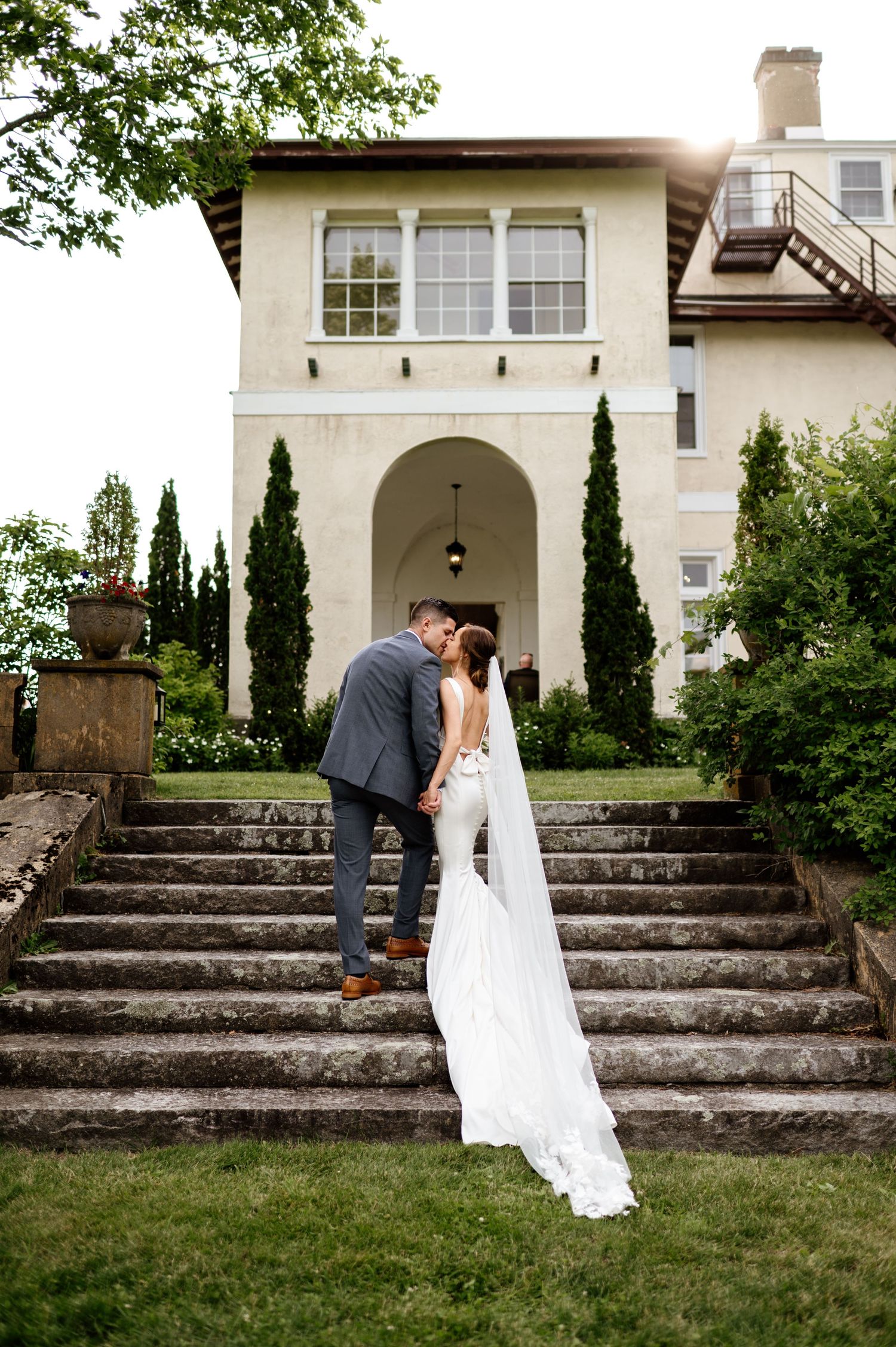 A romantic wedding photo of a couple on stone steps of a Mediterranean-style villa with cypress trees and sunlight.