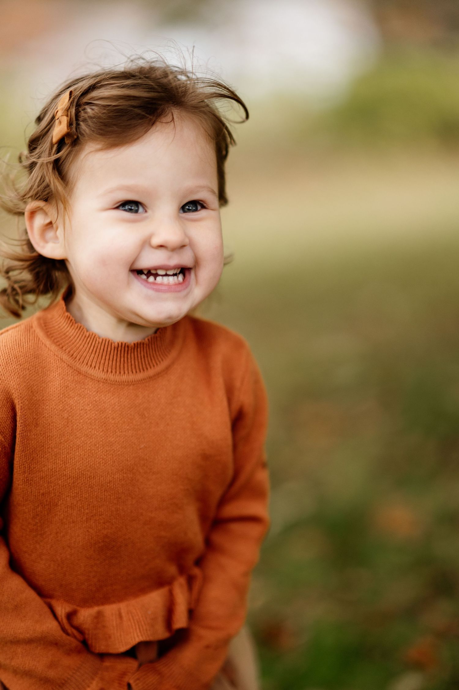 A toddler in a rust-colored sweater smiles brightly outdoors.