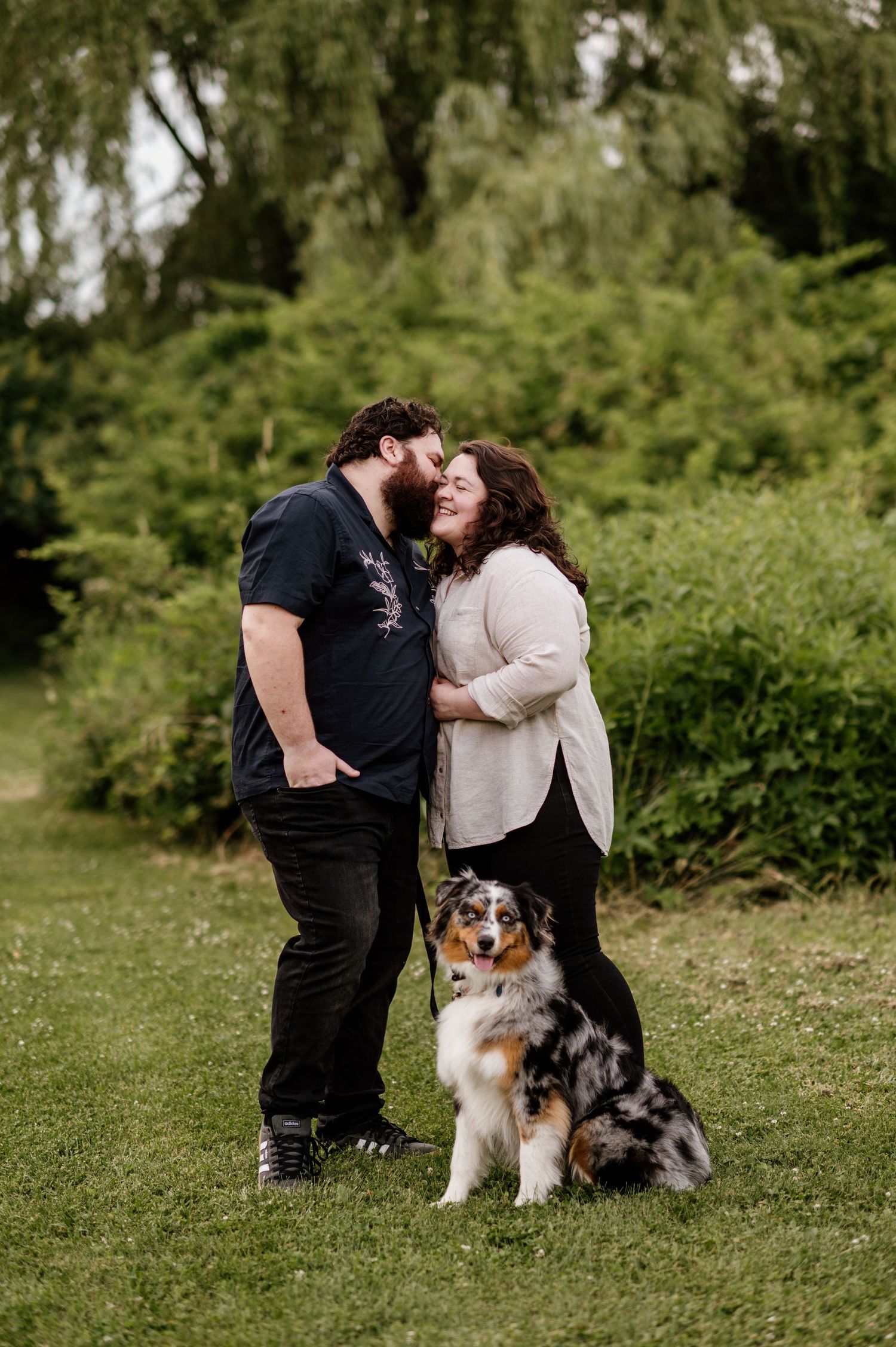 A couple shares an intimate moment in a garden with their Australian Shepherd dog sitting beside them.