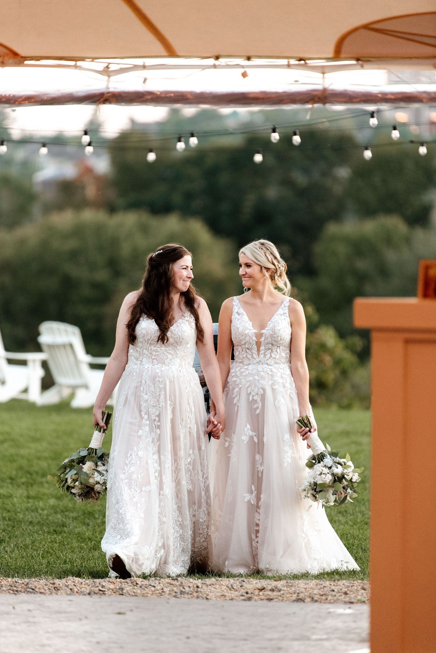 Two people in white wedding dresses walking together outdoors under an orange umbrella with string lights.