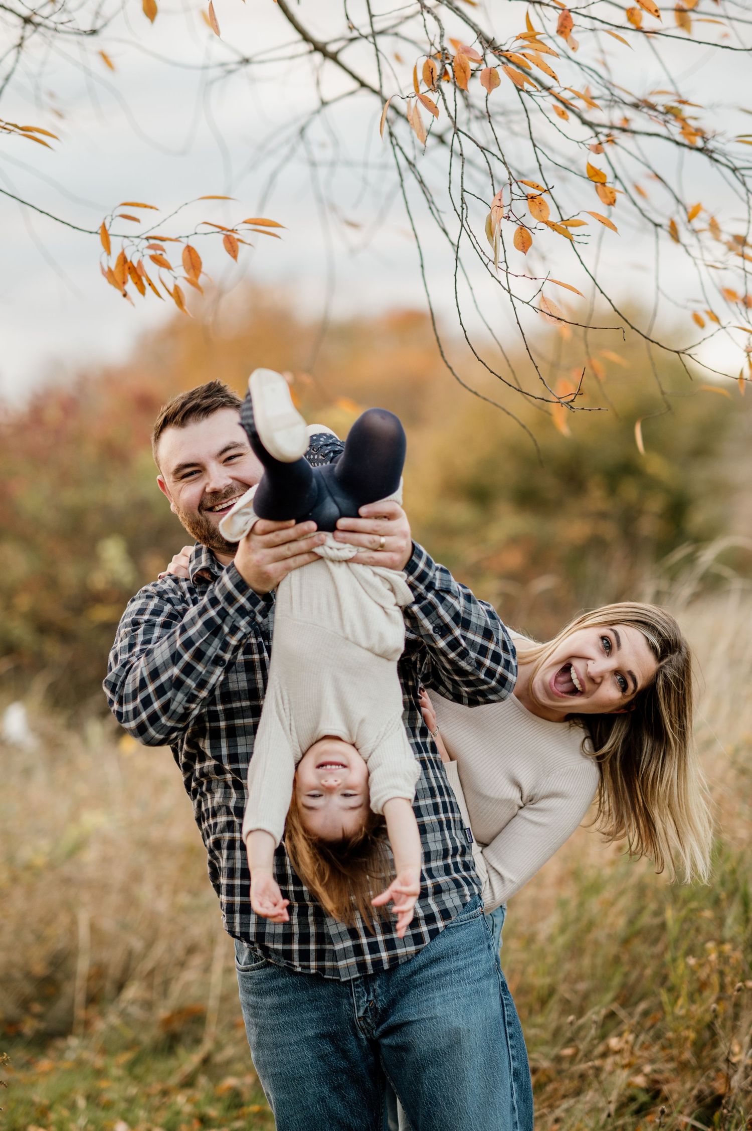 Playful couple laughing and embracing in an autumn field with orange leaves overhead.