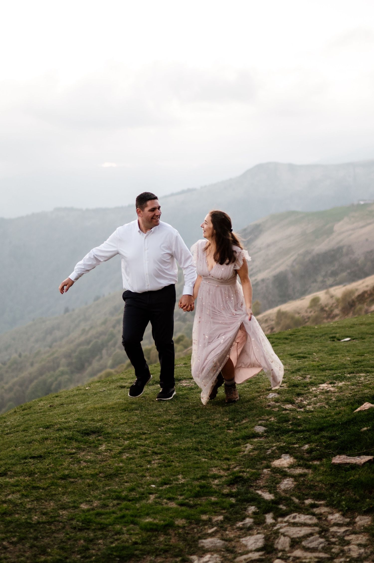 Couple in wedding attire playfully running on mountain hillside with scenic views.