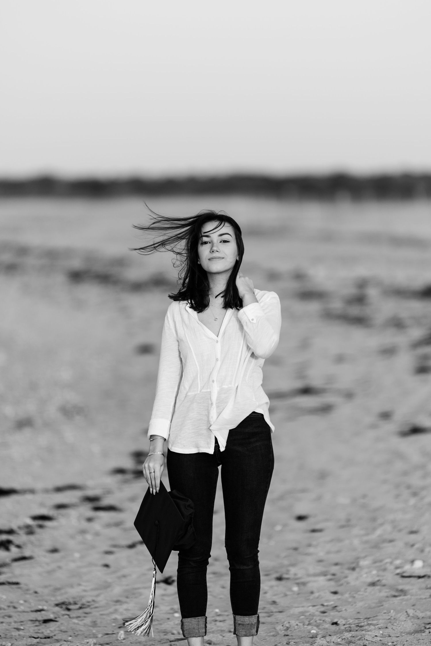 A person in a white button-down shirt and dark pants stands on a beach with windswept hair in black and white.