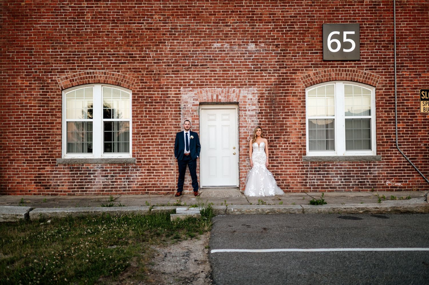 Wedding couple poses against industrial brick wall backdrop with vintage windows and building number.