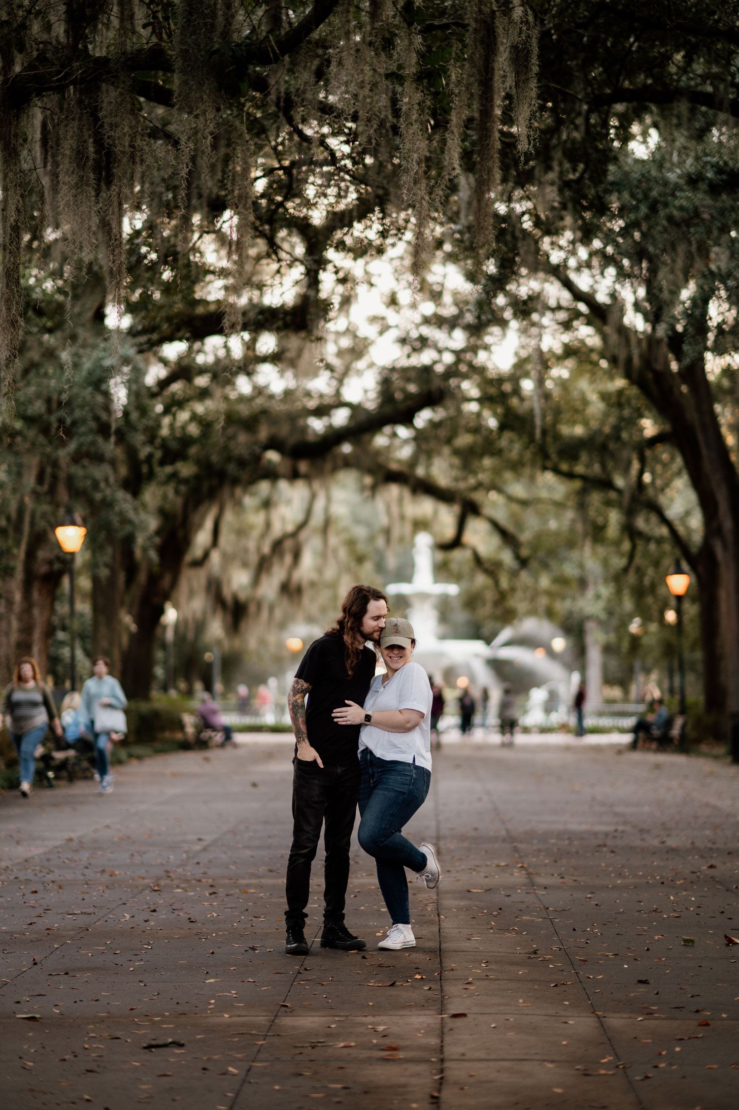 A couple shares a romantic moment beneath Spanish moss-draped trees in a park setting at sunset.